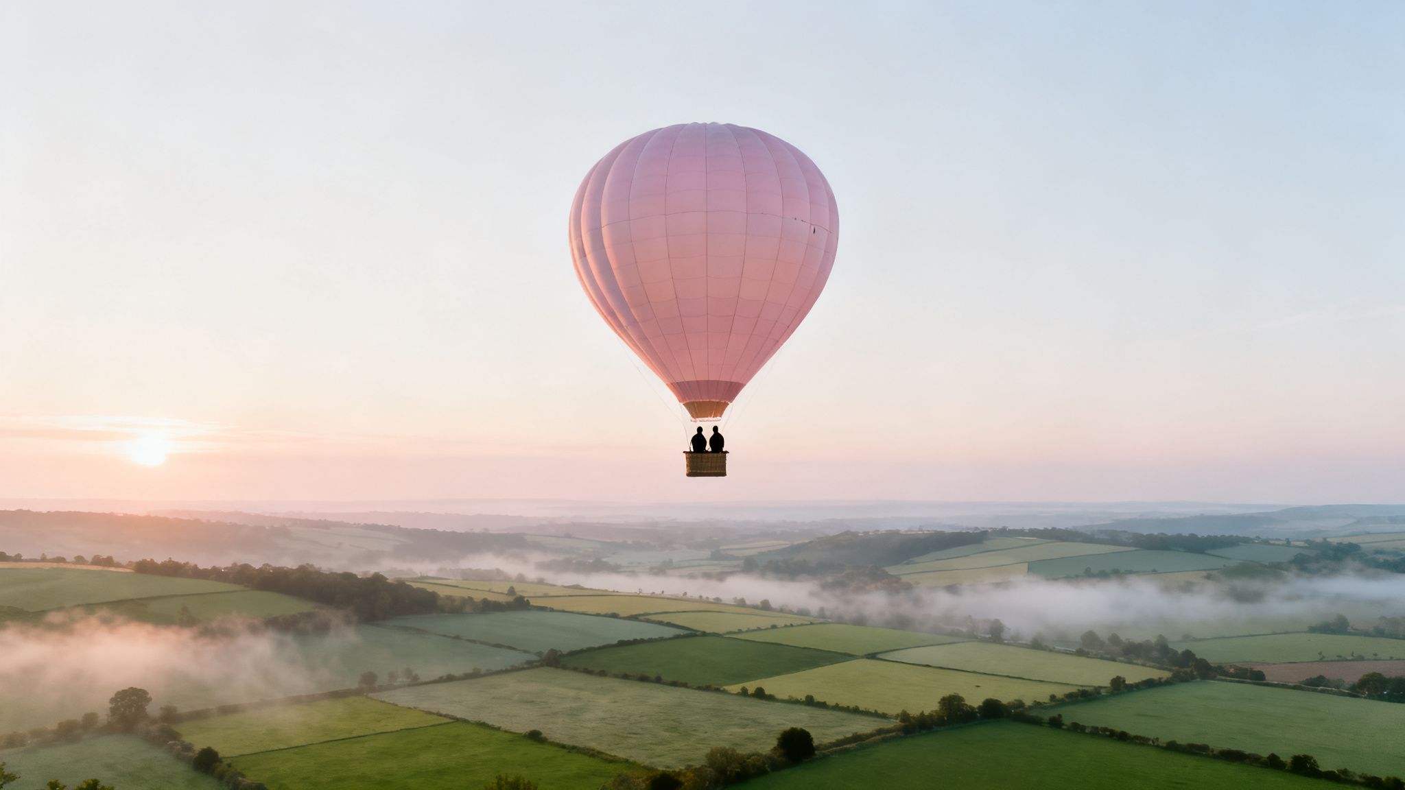 Pink hot air balloon with two people soaring over misty green fields at sunrise.
