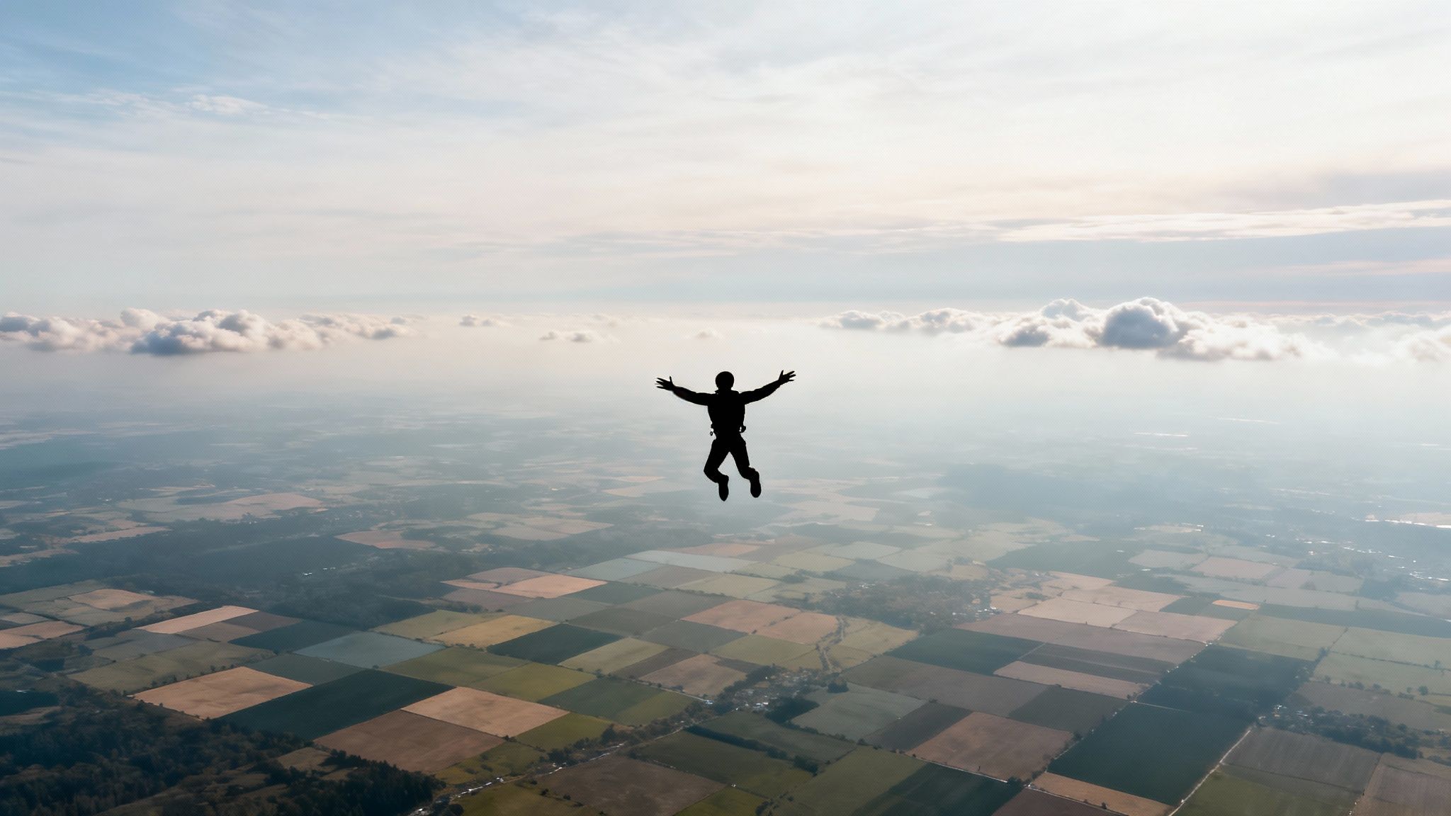 A silhouette of a person skydiving with arms and legs spread, high above a patchwork of fields and clouds.