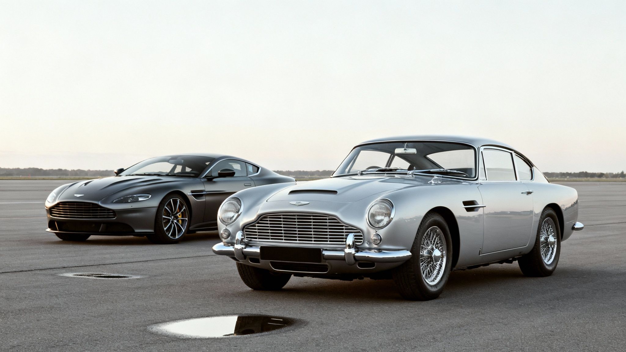 A classic silver Aston Martin DB5 and a modern dark grey Vantage parked on an airfield.