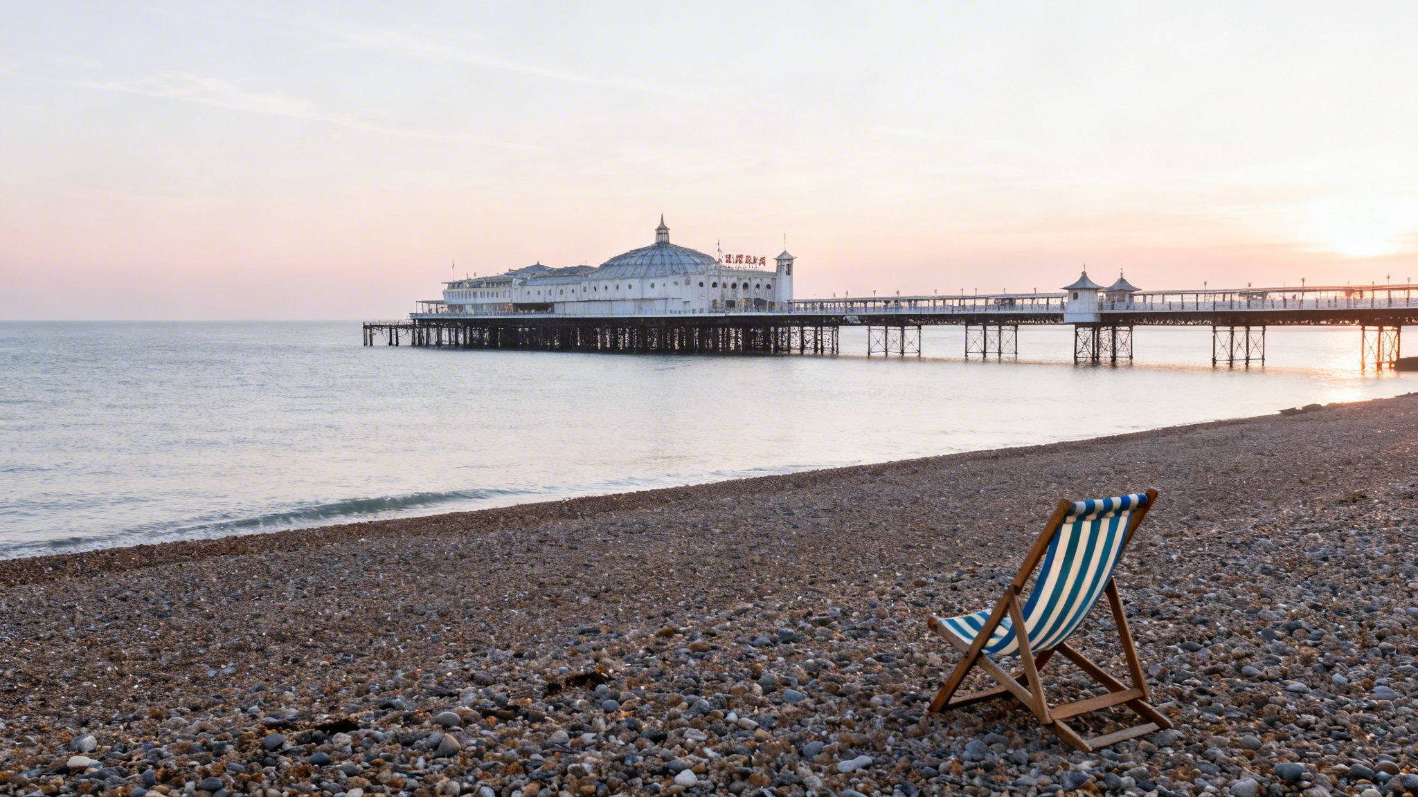 A serene pebble beach with a deckchair facing a historic pier at sunset, UK.
