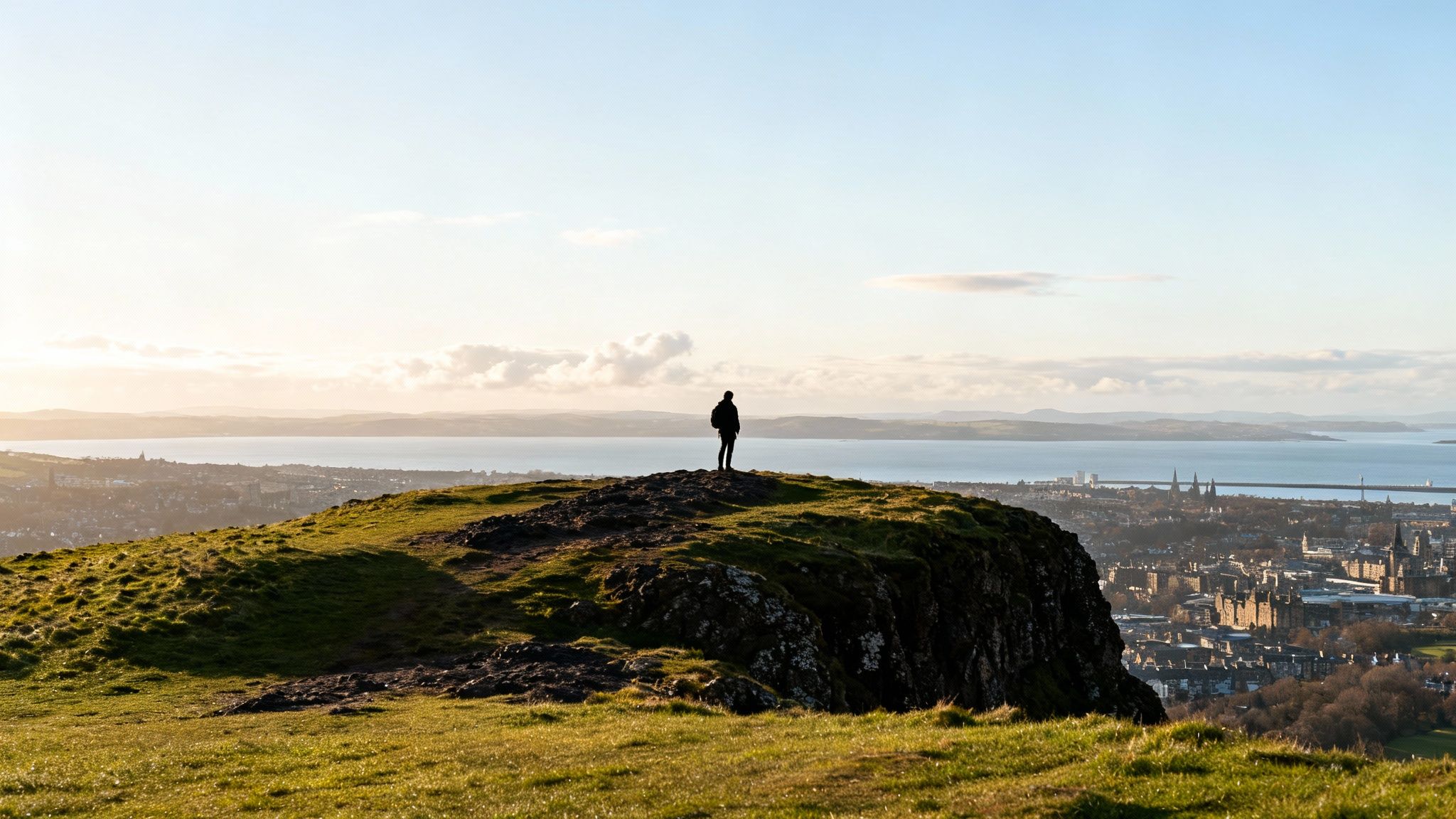 A lone person stands atop a grassy hill overlooking the city of Edinburgh and a sea loch at sunset.