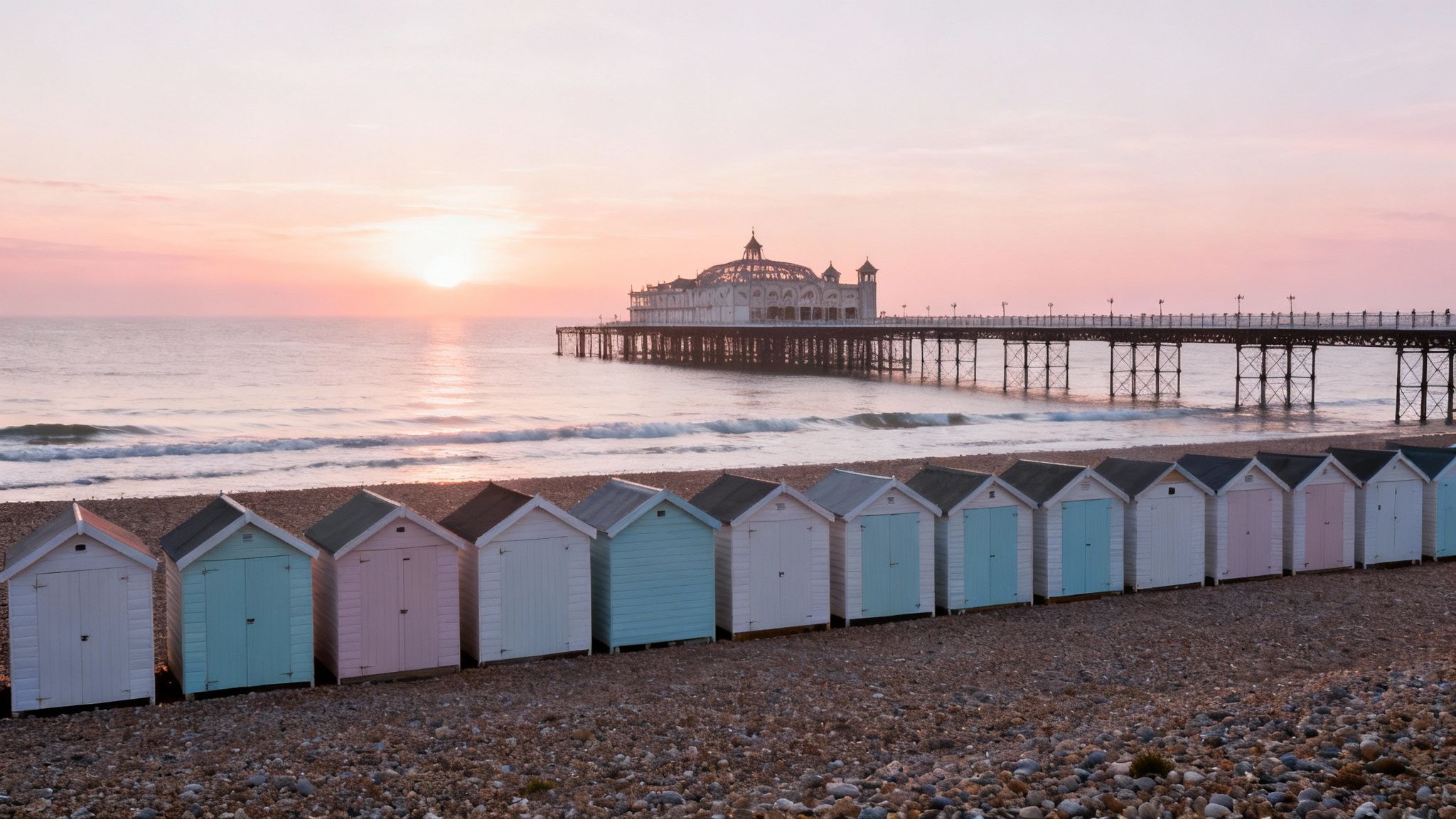 A scenic sunset over the sea with a pier and colorful beach huts on a pebble beach.