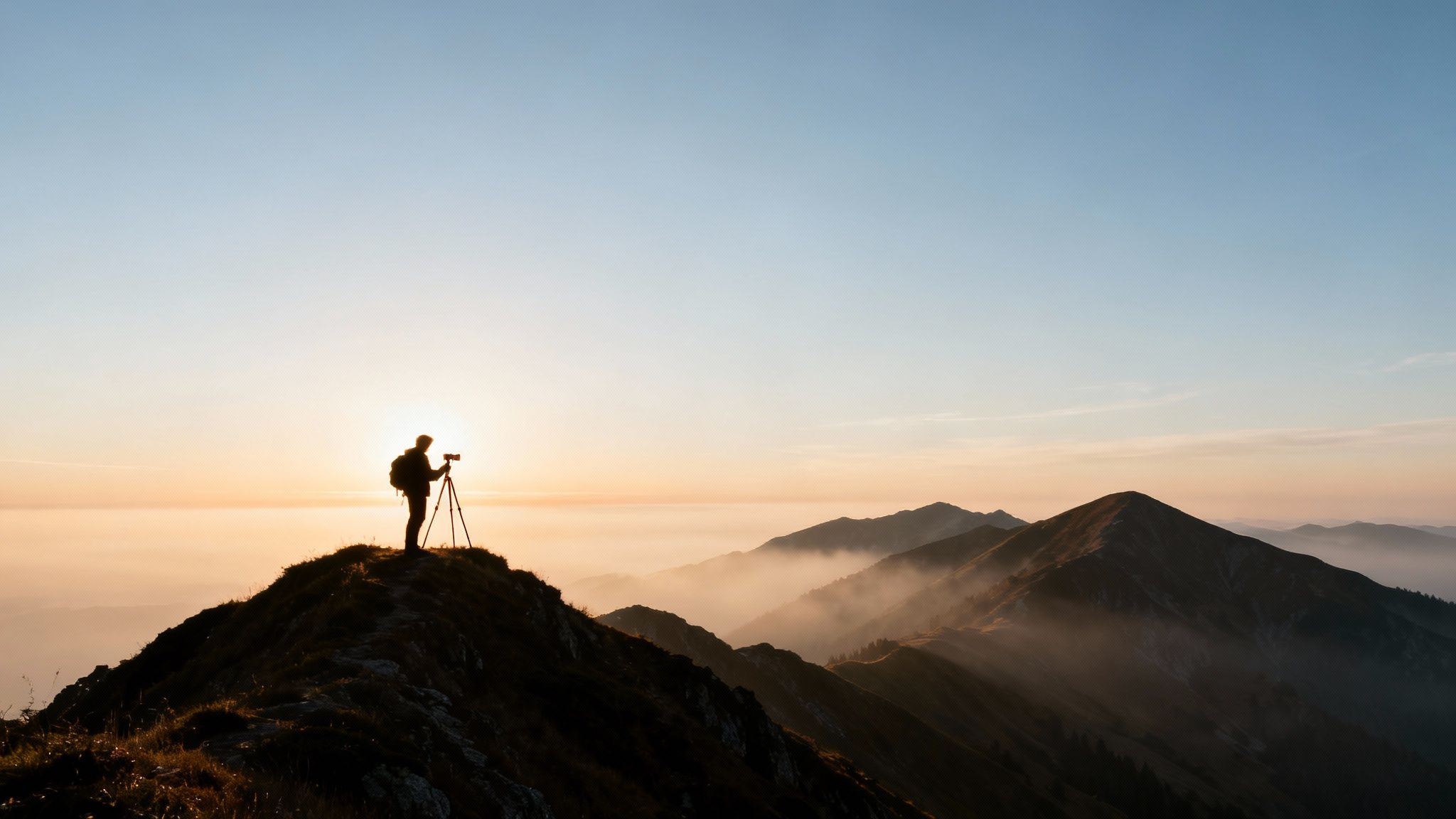 A silhouette of a photographer with a camera on a tripod on a mountain ridge at sunrise over foggy valleys.