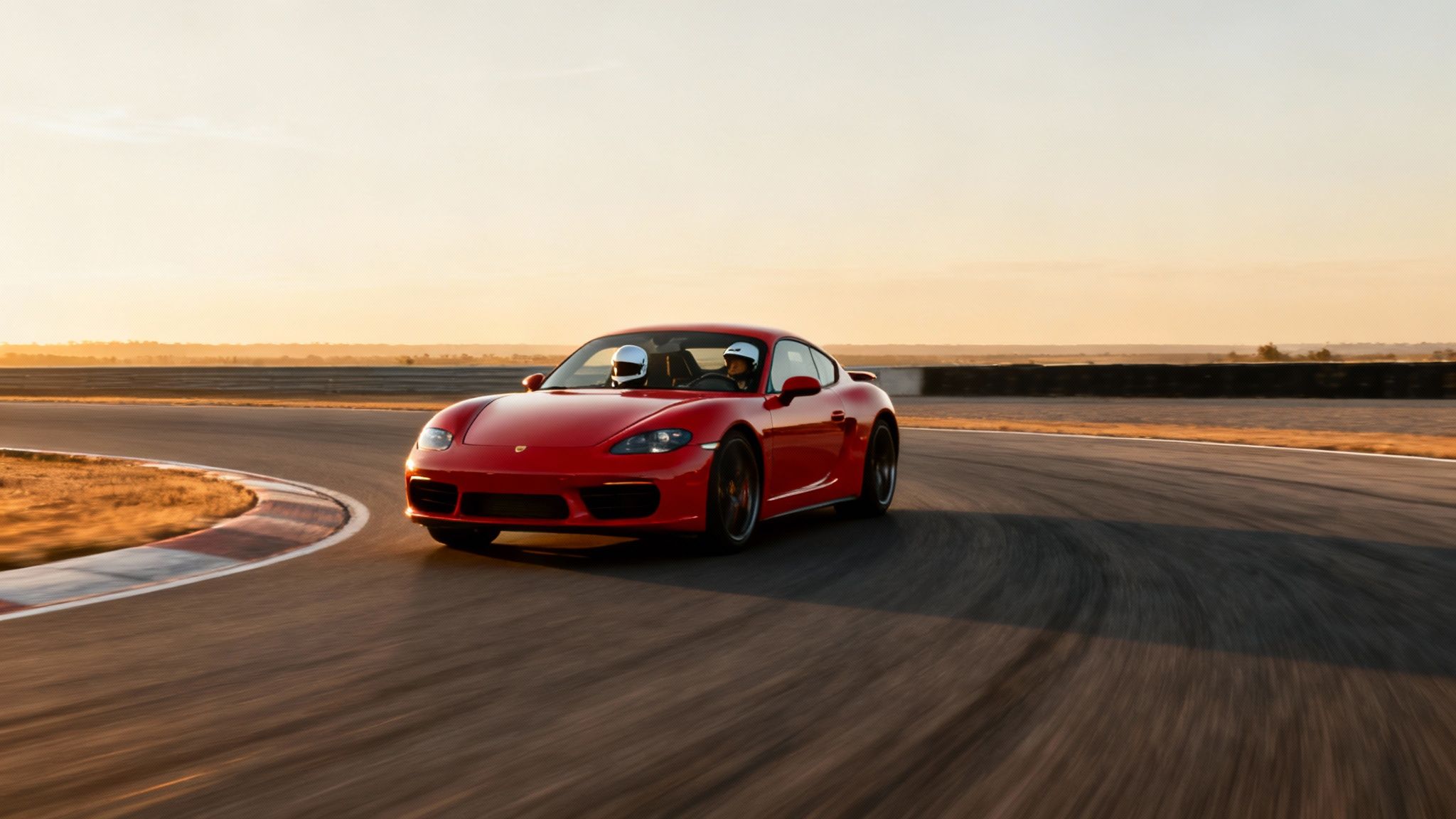 Red Porsche sports car with two drivers in helmets racing on a track at sunset.