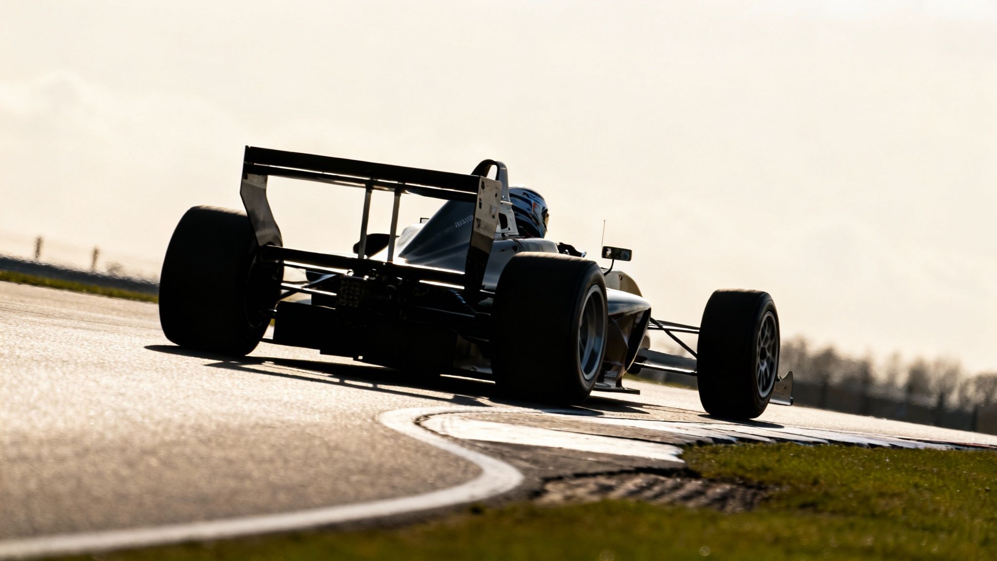 Rear view of a formula race car with a driver's helmet visible, on a bright track corner.