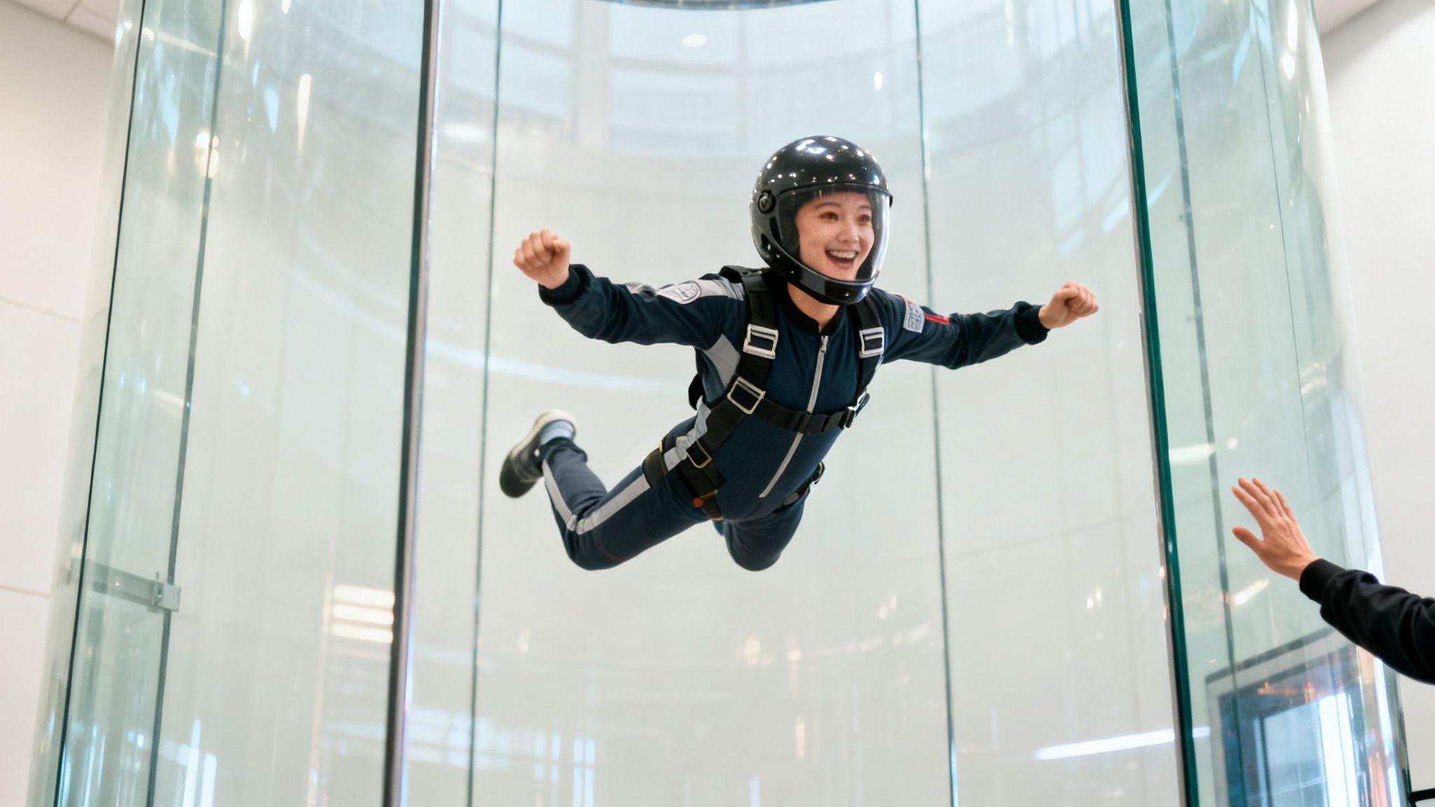 A smiling woman in a helmet and jumpsuit experiences indoor skydiving in a glass wind tunnel.