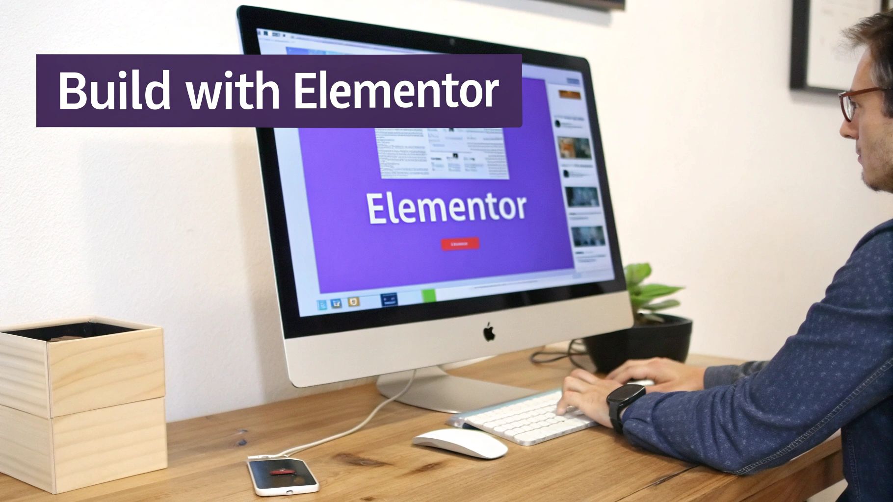 A man works on an iMac displaying Elementor website builder, with a keyboard and mouse on a wooden desk.