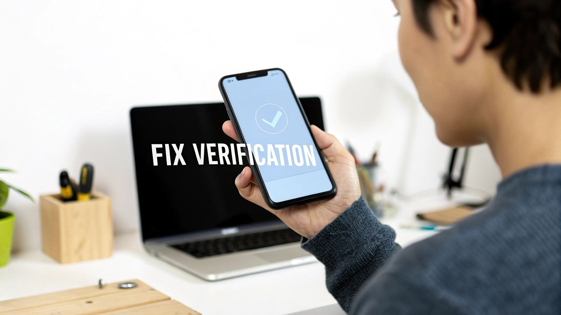 A person holds a smartphone displaying a successful verification checkmark, with a laptop in the background on a desk.