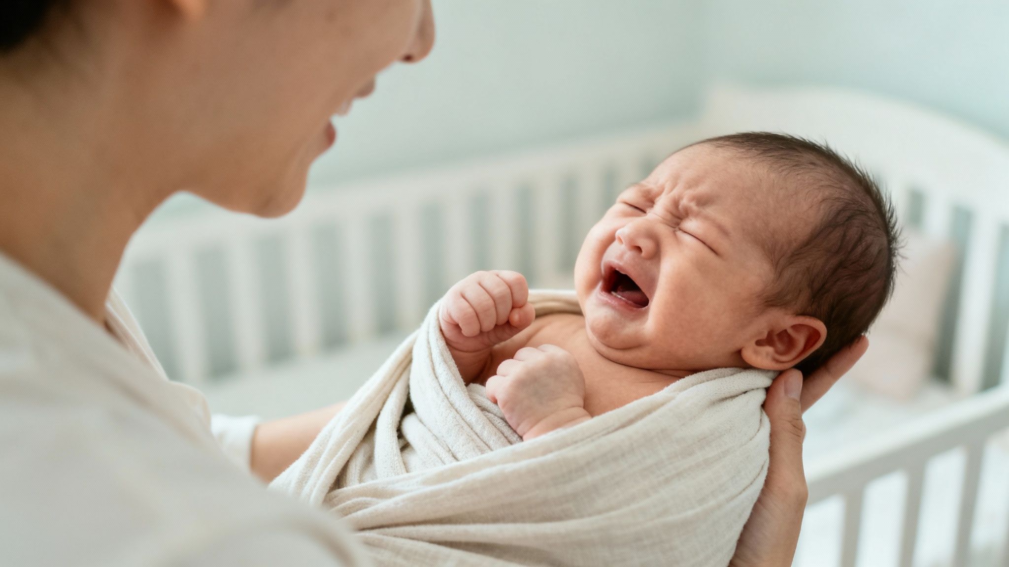 A parent comforts a swaddled baby who is crying loudly in a bright nursery.