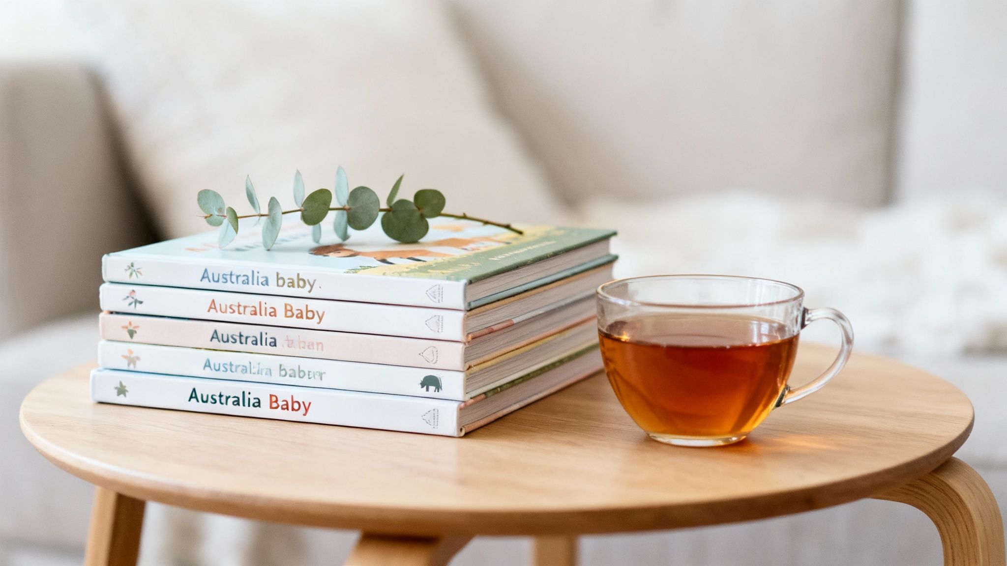 A stack of 'Australia Baby' children's books with an eucalyptus branch and a cup of tea on a wooden table.