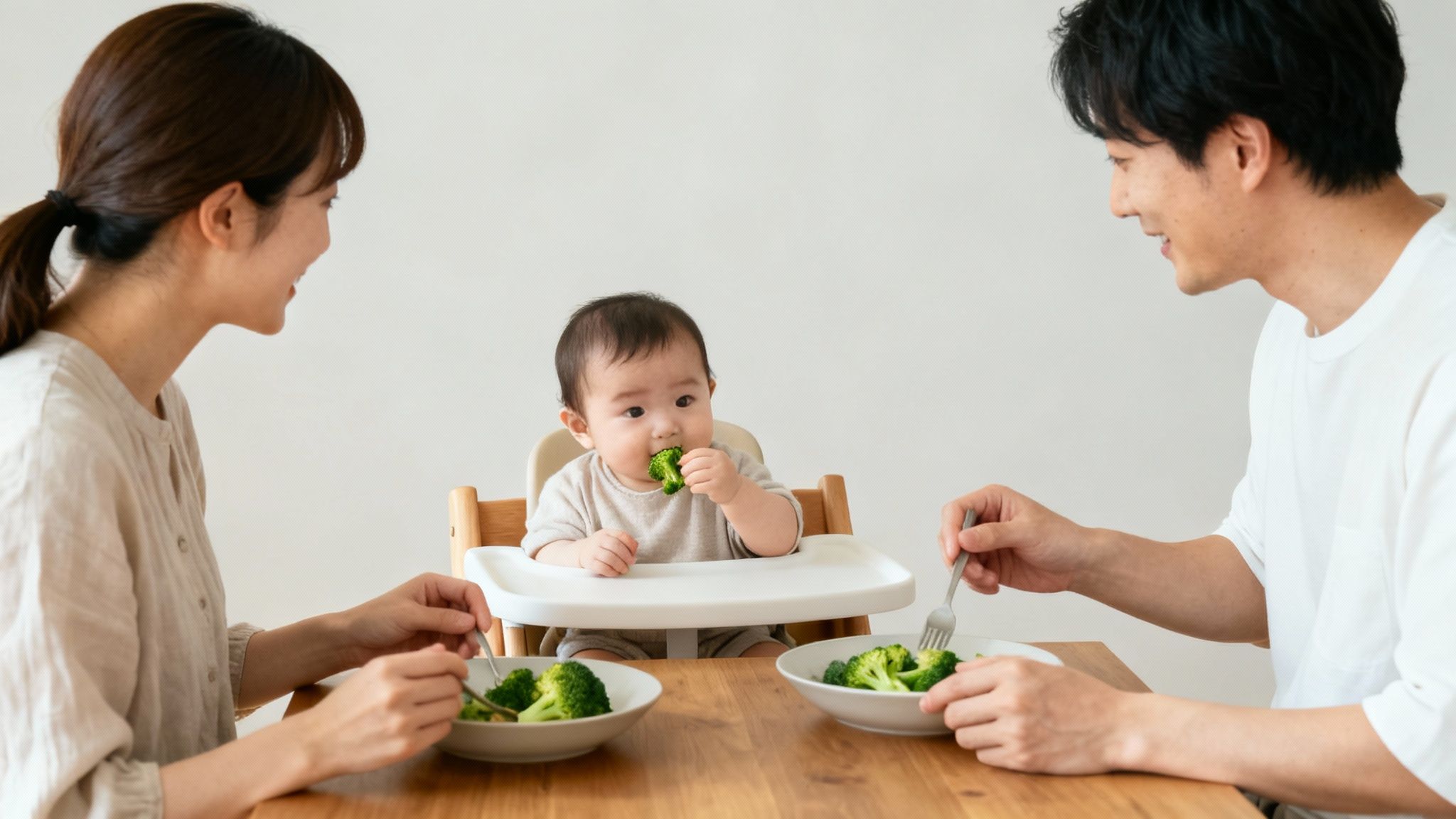 A happy family of three sharing a healthy meal, with baby in high chair eating broccoli.
