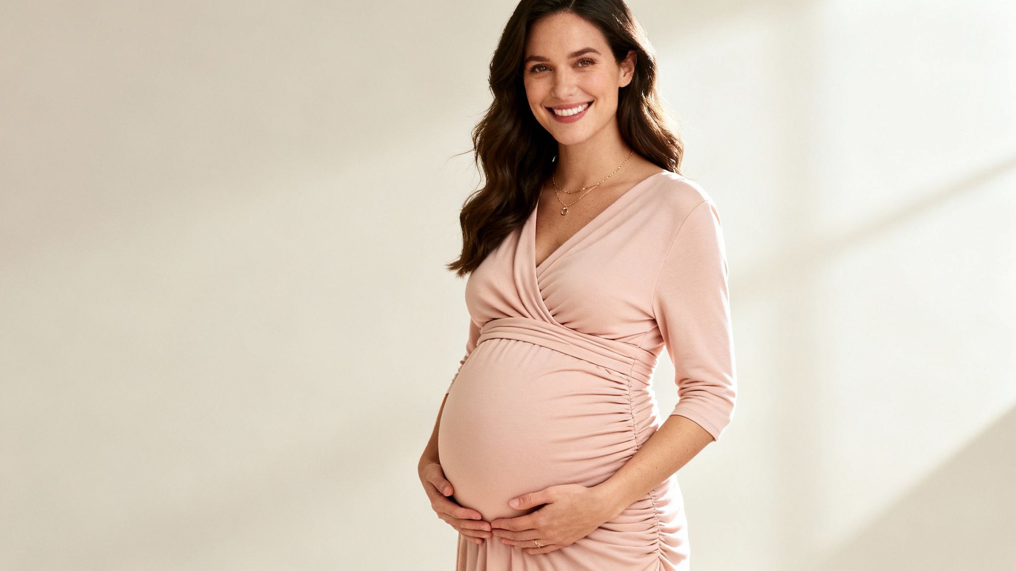 A smiling pregnant woman in a pink wrap dress gently holding her baby bump.