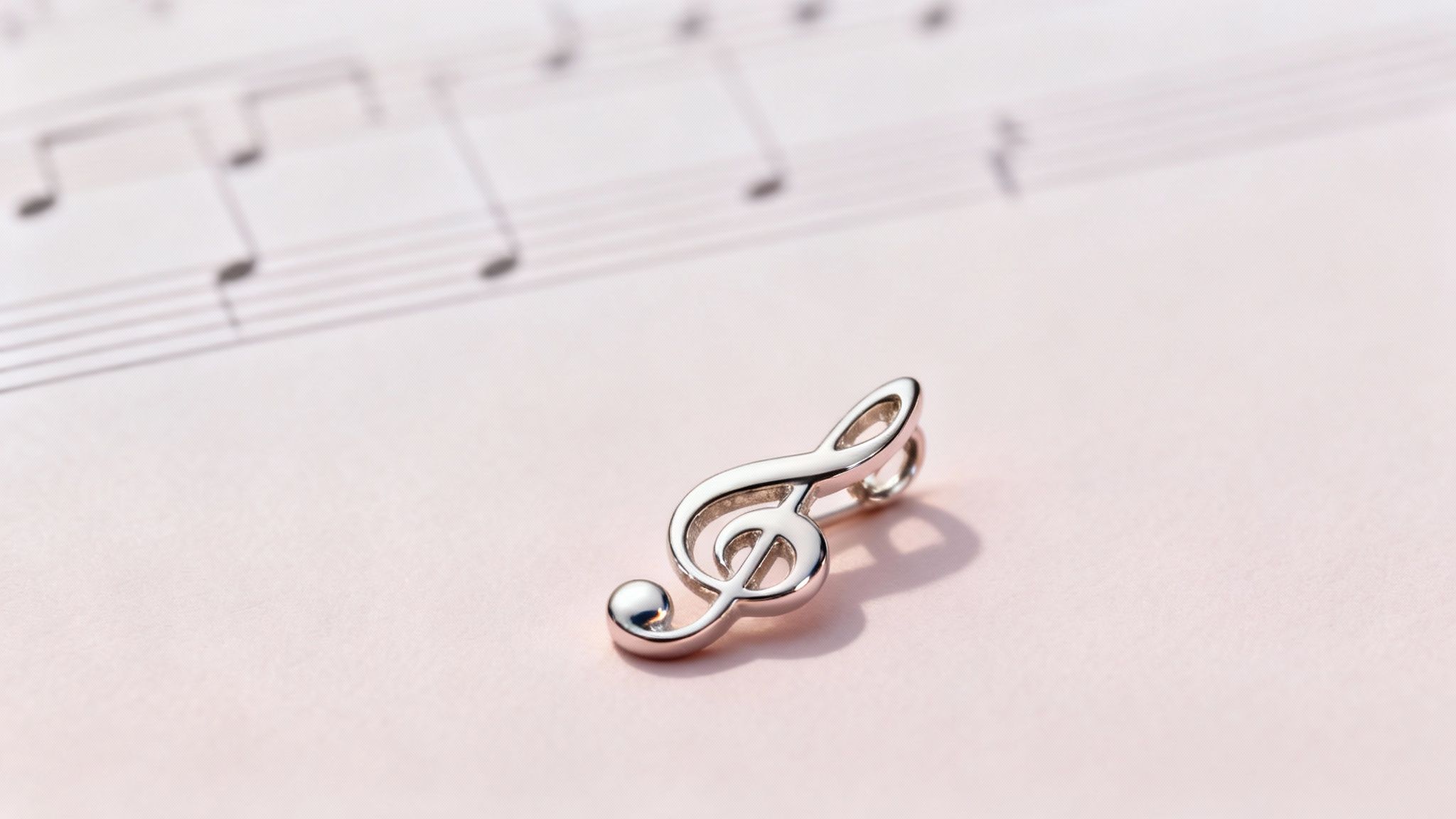 A shiny silver treble clef pendant rests on a light pink surface with blurred sheet music in the background.