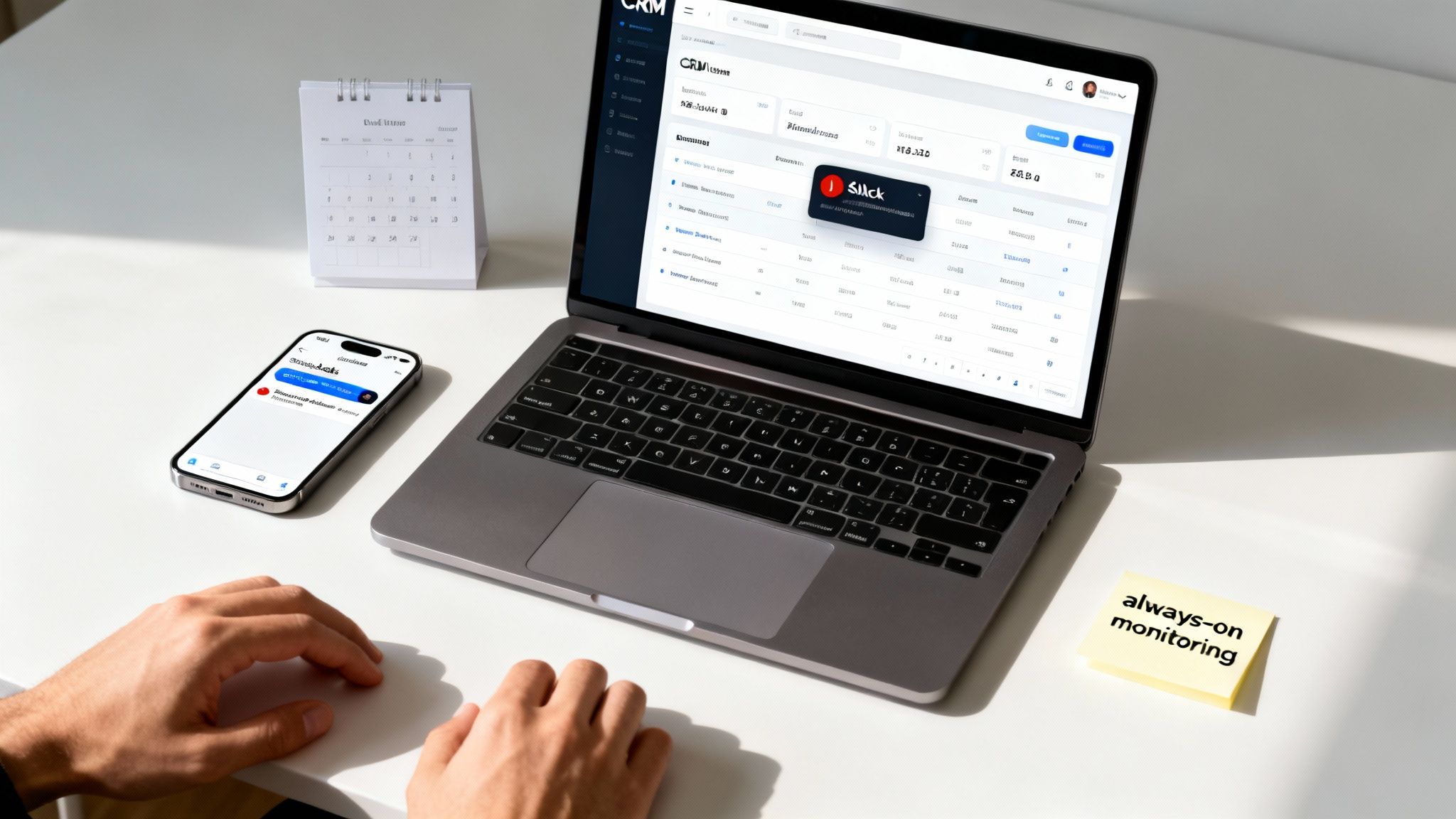 Person's hands on a white desk, monitoring a CRM system on a laptop with a phone and calendar.