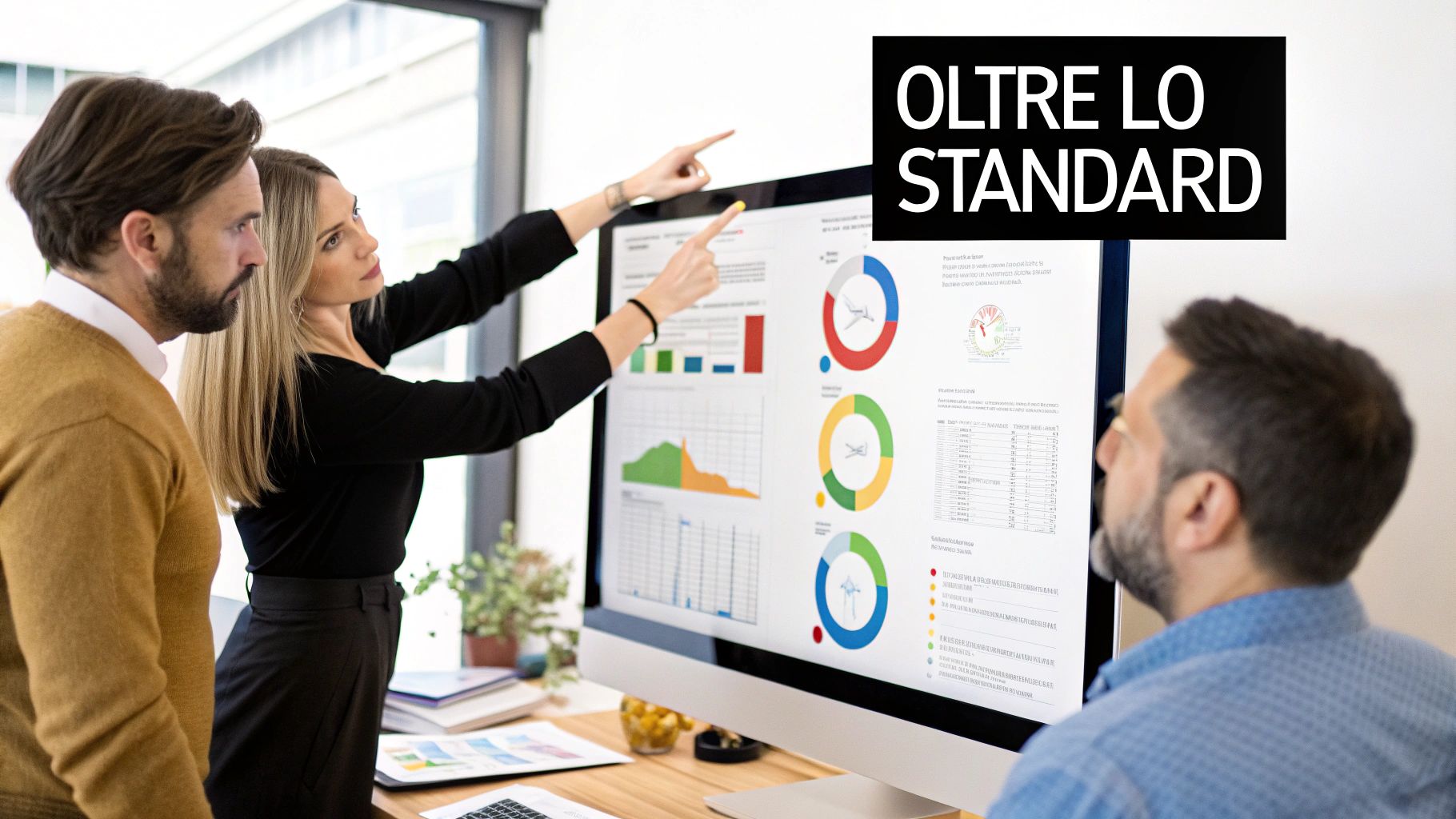 Tre professionisti in ufficio esaminano grafici e dati su un monitor, con una donna che indica lo schermo. 'OLTRE LO STANDARD' in evidenza.