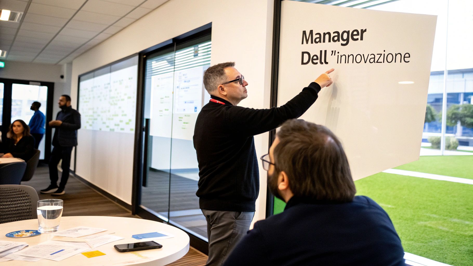 Un uomo spiega, indicando una lavagna con il testo "Manager Dell "innovazione"", durante una riunione in ufficio.