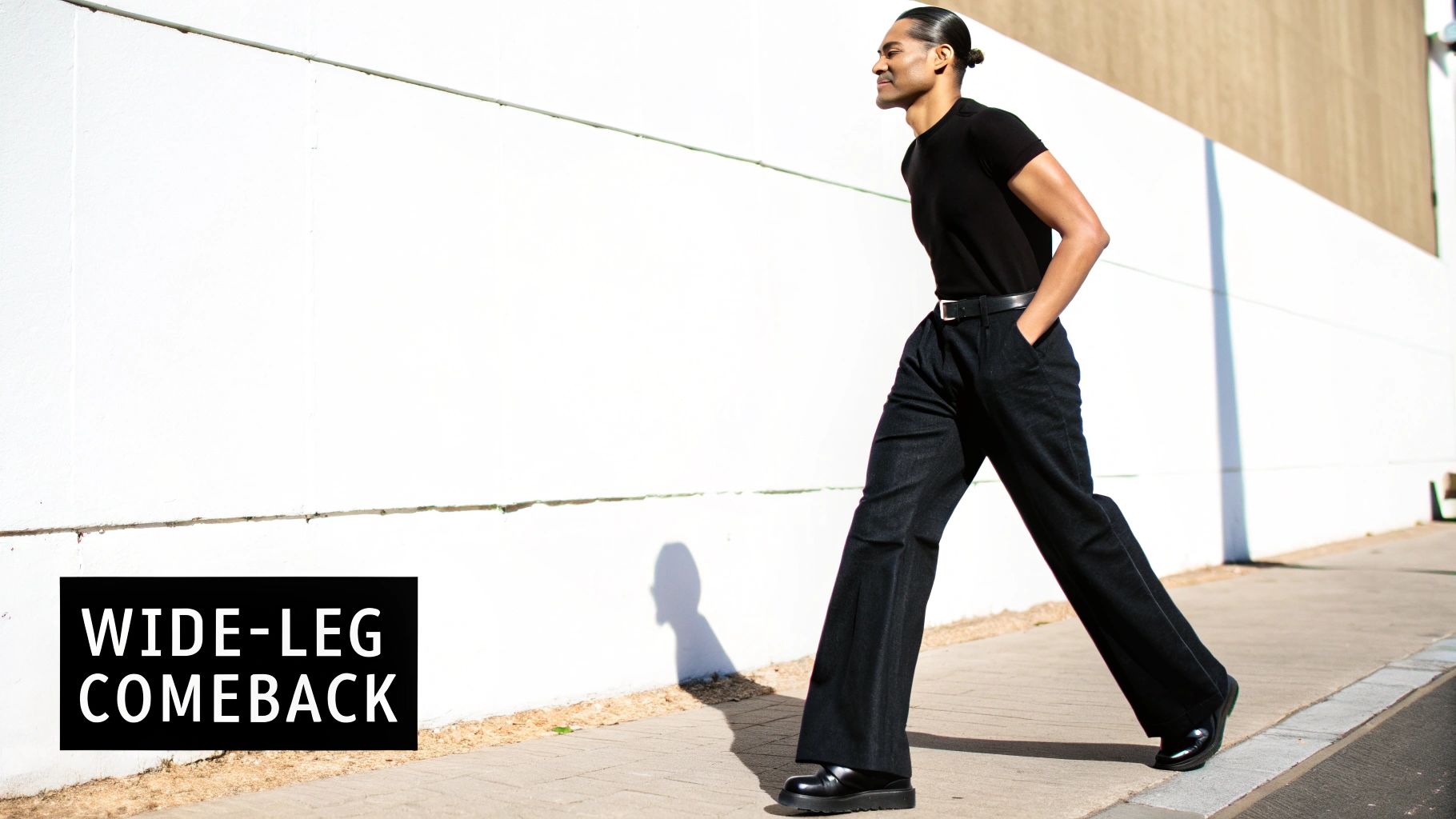 A stylish man in a black t-shirt and wide-leg dark pants walks on a sidewalk.