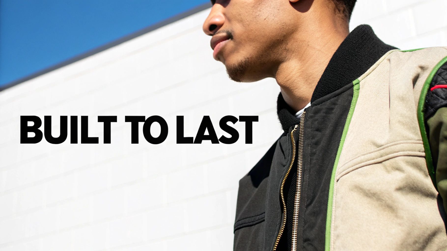 A man in a multi-colored bomber jacket with the text "BUILT TO LAST" against a white wall and blue sky.
