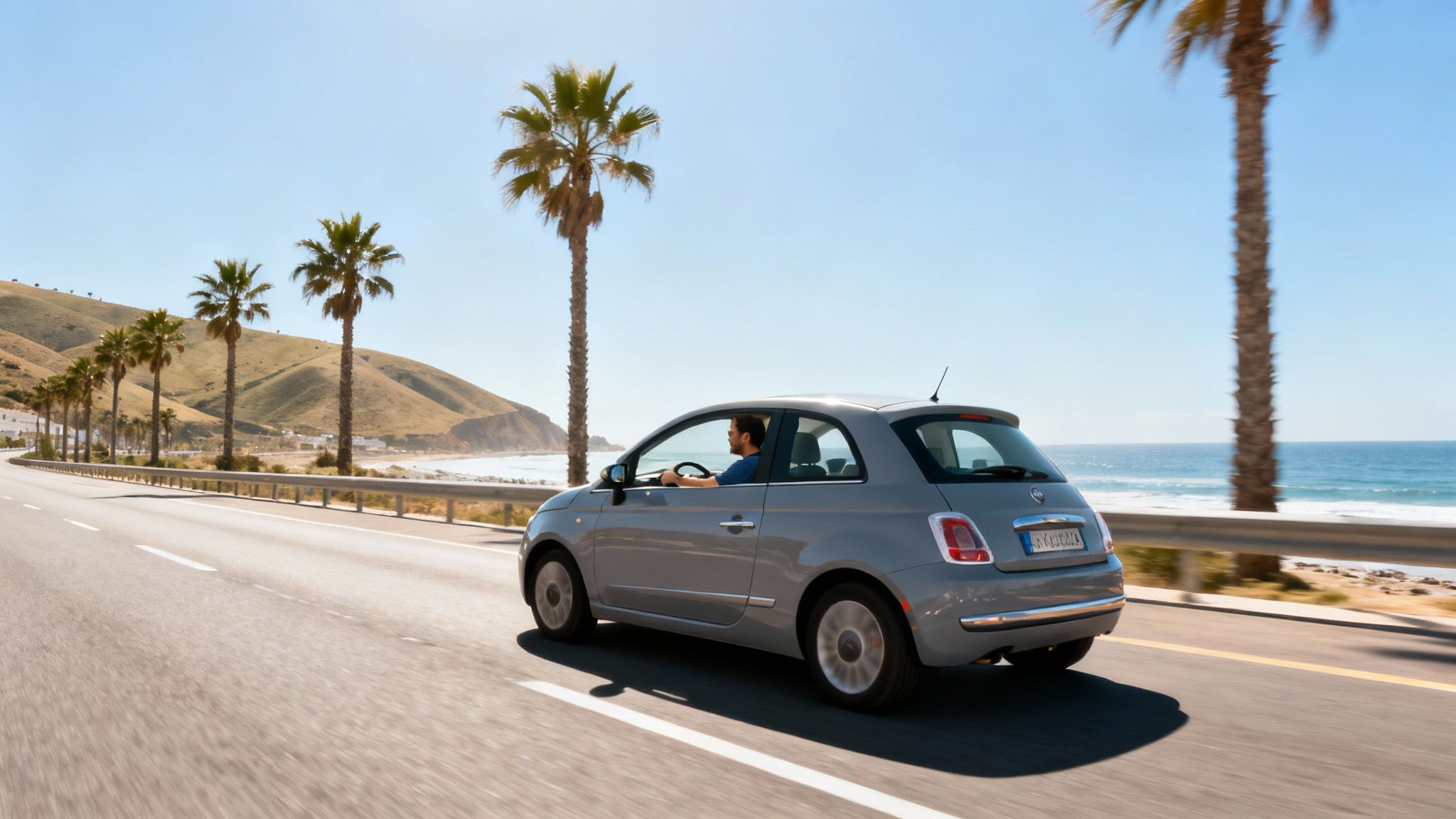 Une voiture grise roule sur une route côtière ensoleillée avec des palmiers et la mer bleue en arrière-plan.