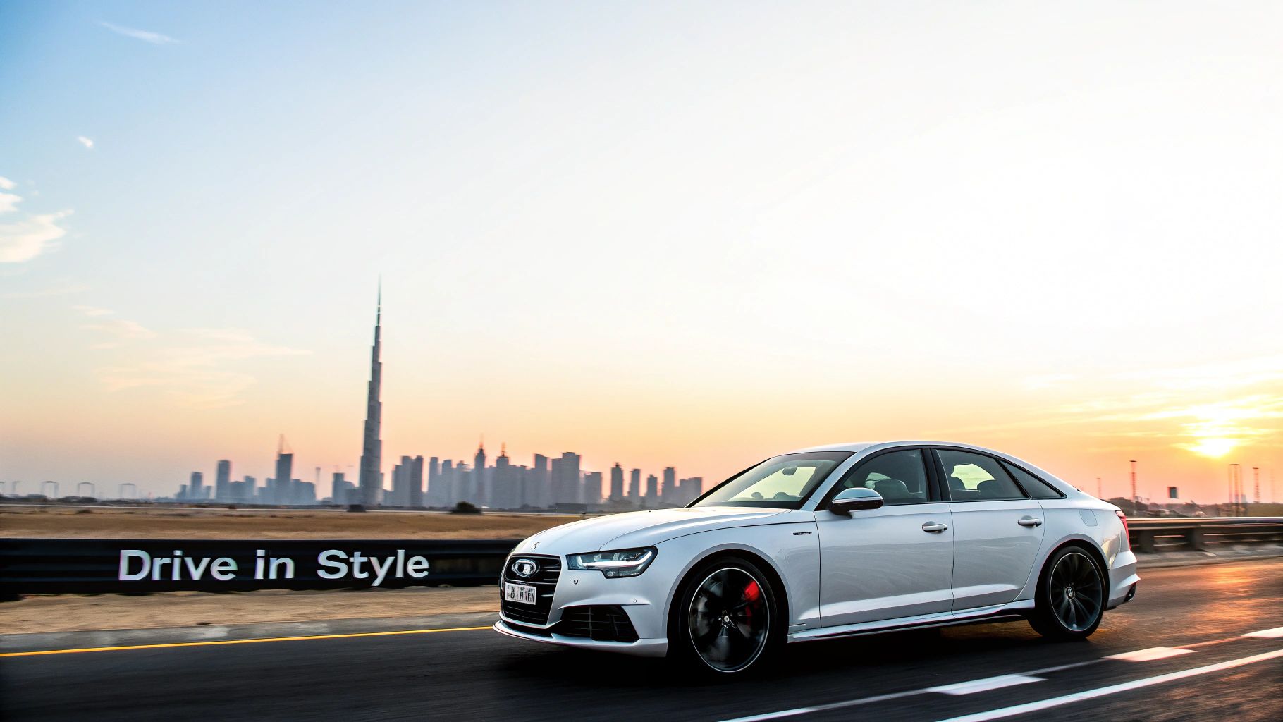 White Audi car driving on a road with Dubai skyline and Burj Khalifa at sunset.