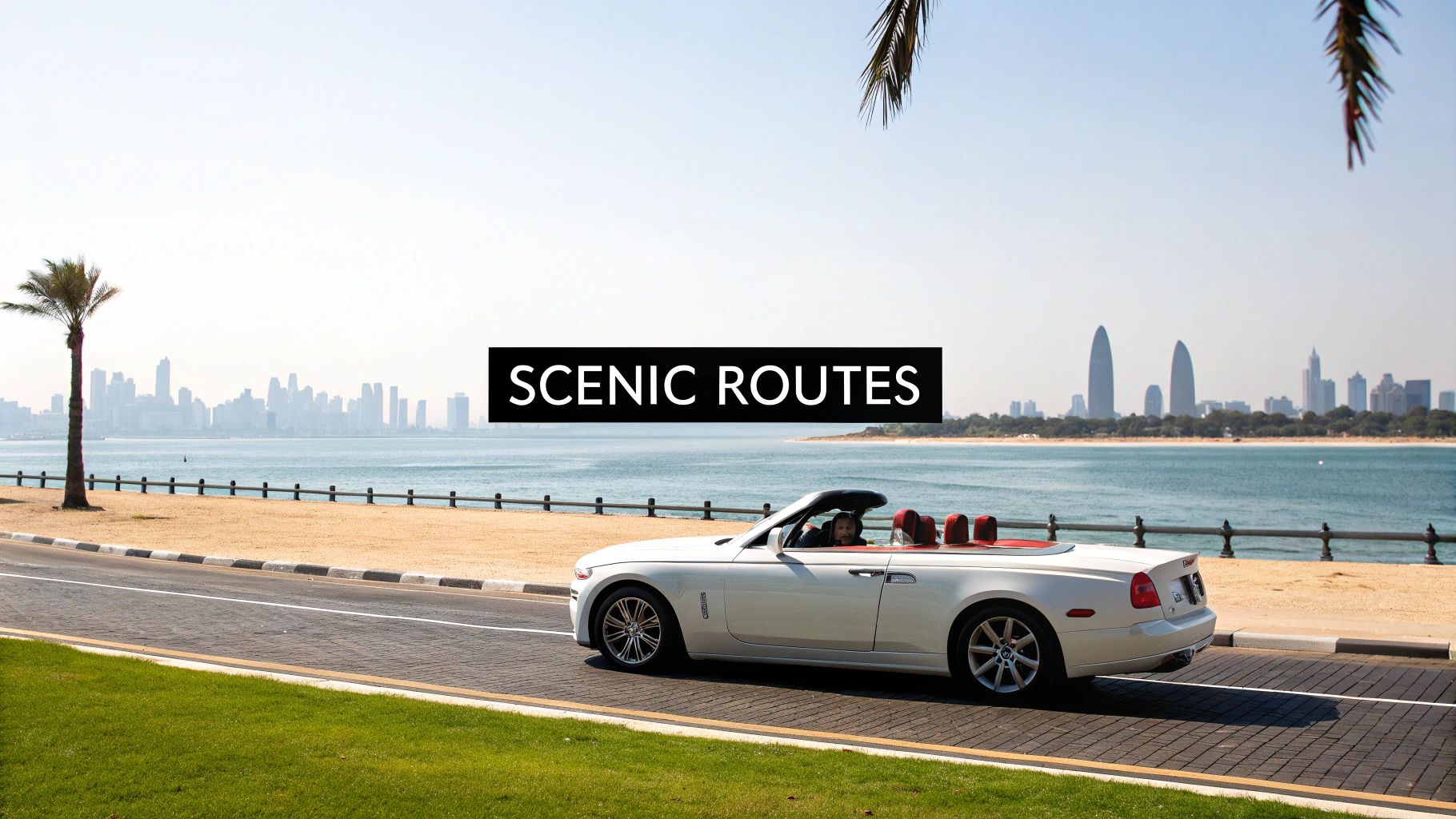 A white Rolls Royce Dawn convertible on a scenic coastal road with a city skyline and ocean views.