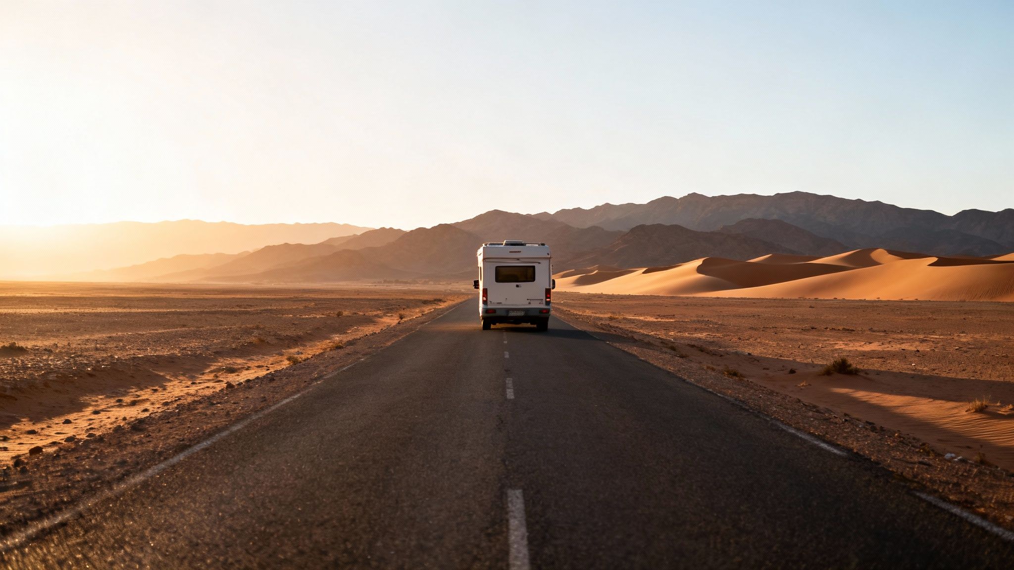 Camping-car blanc sur une route déserte. Paysage marocain avec montagnes et dunes de sable au coucher du soleil.