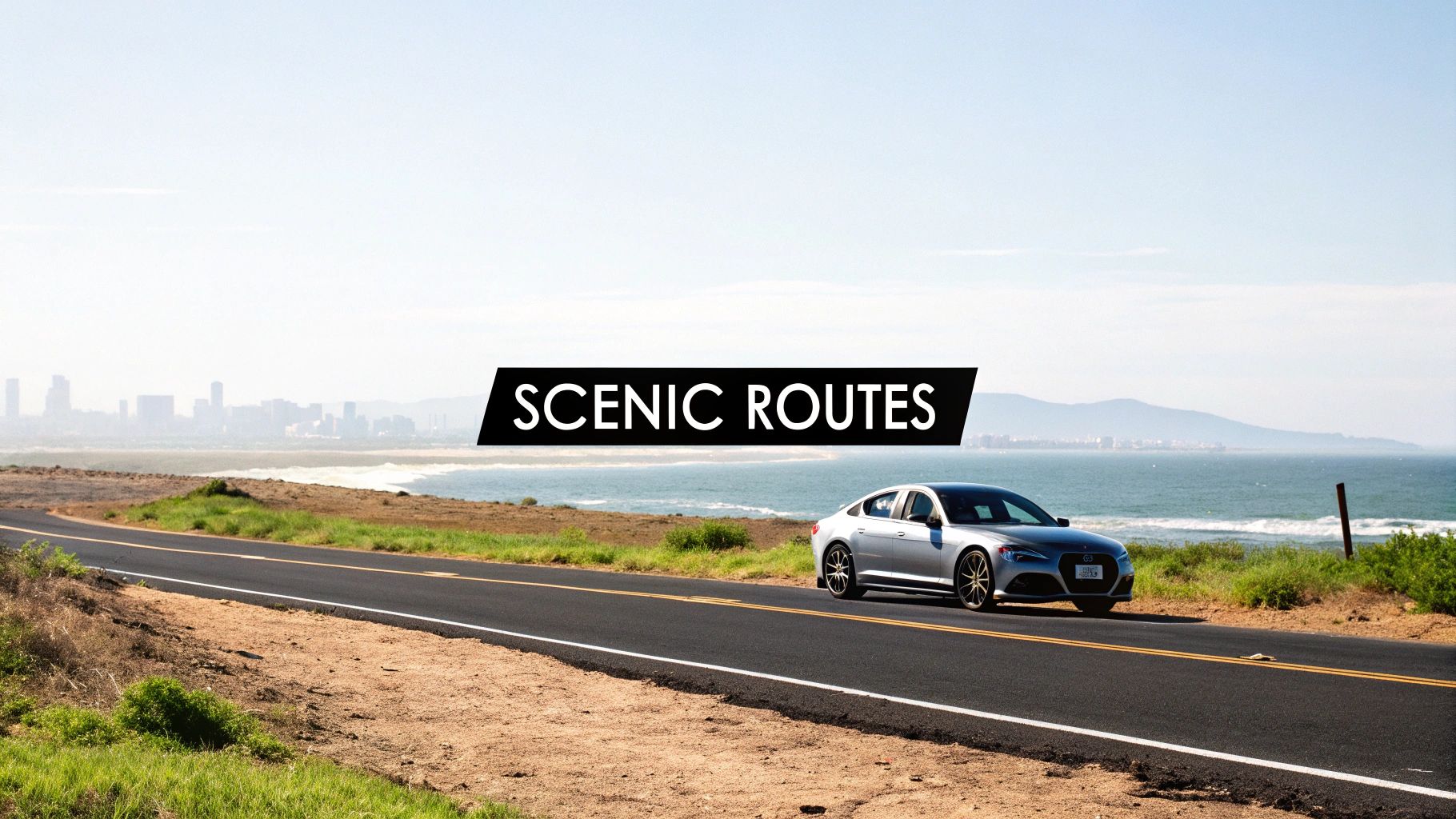 A silver car drives along a scenic coastal route, with the ocean and a city skyline in the background.