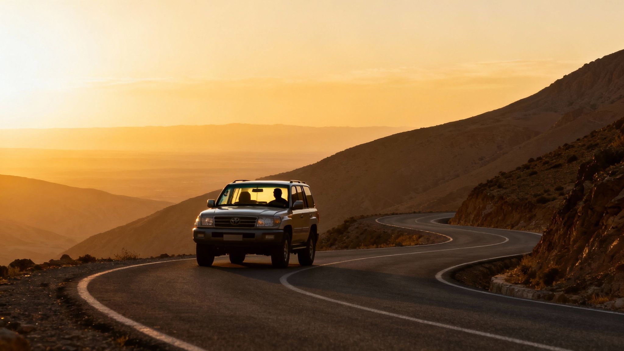 Un SUV gris roule sur une route de montagne sinueuse au coucher du soleil avec un ciel doré.
