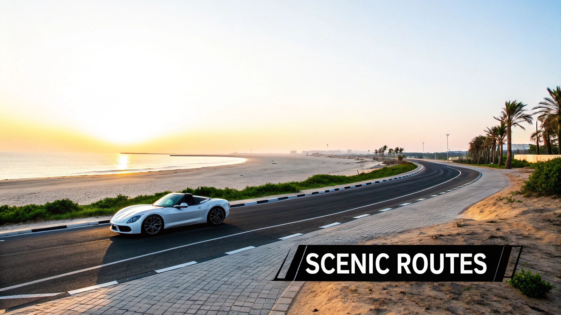 A white convertible sports car drives on a scenic coastal road beside a beach at sunset.
