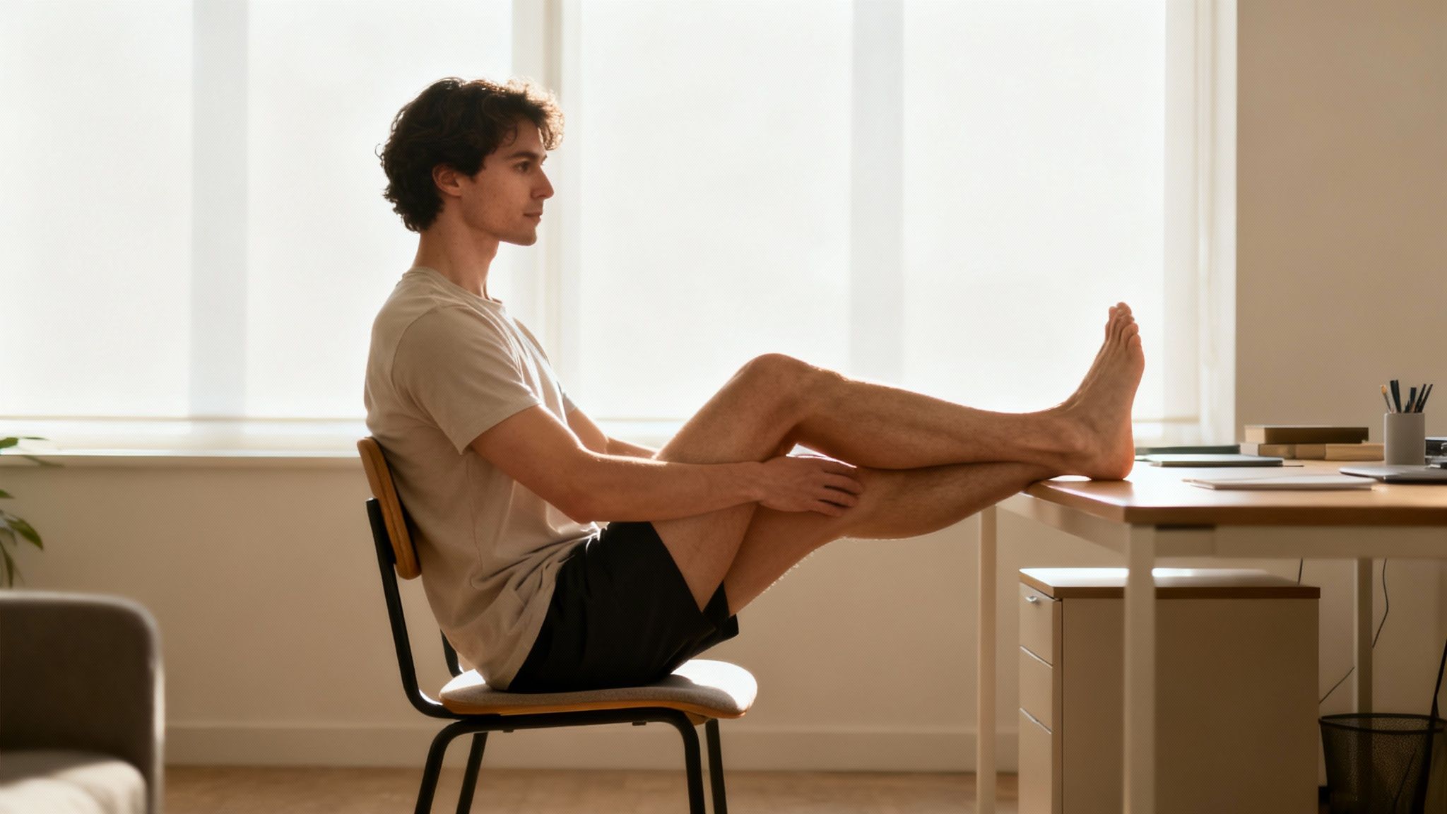 Relaxed young man in a t-shirt and shorts, feet up on a desk, near a bright window.