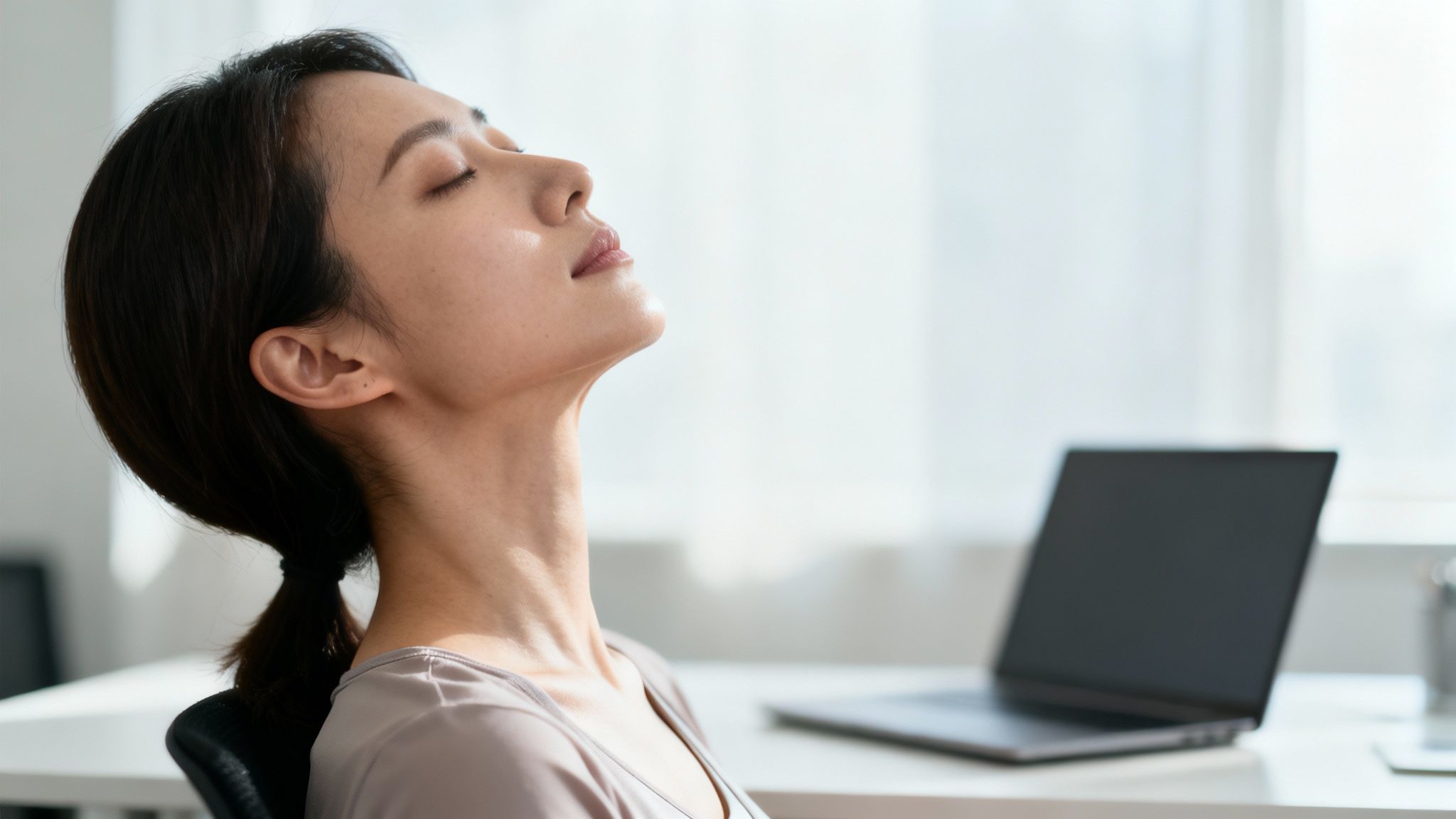 A woman with eyes closed, tilting her head back, relaxing at her desk.