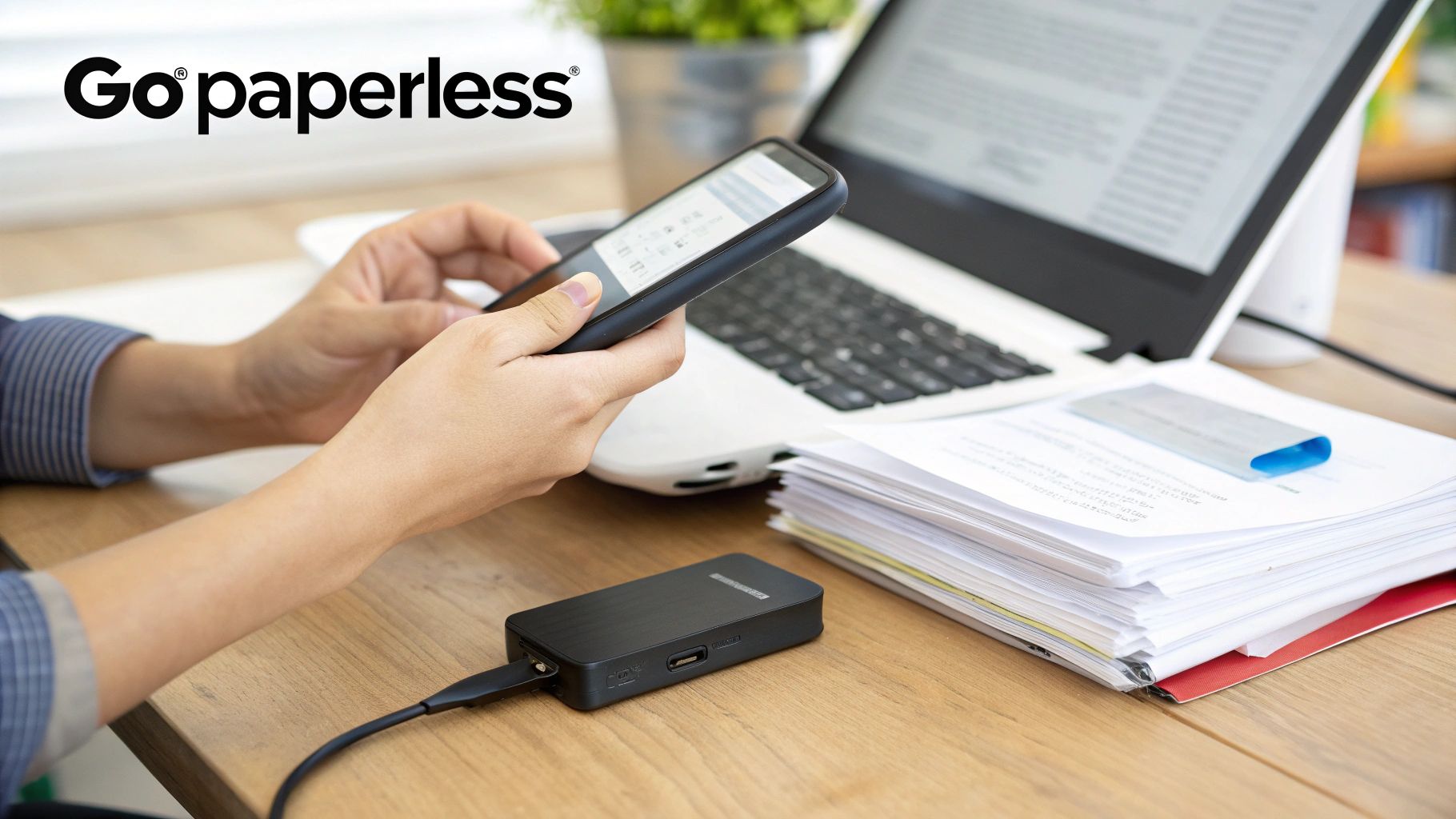 Person using a smartphone and laptop on a wooden desk with a portable device and stack of papers, promoting a paperless workflow.