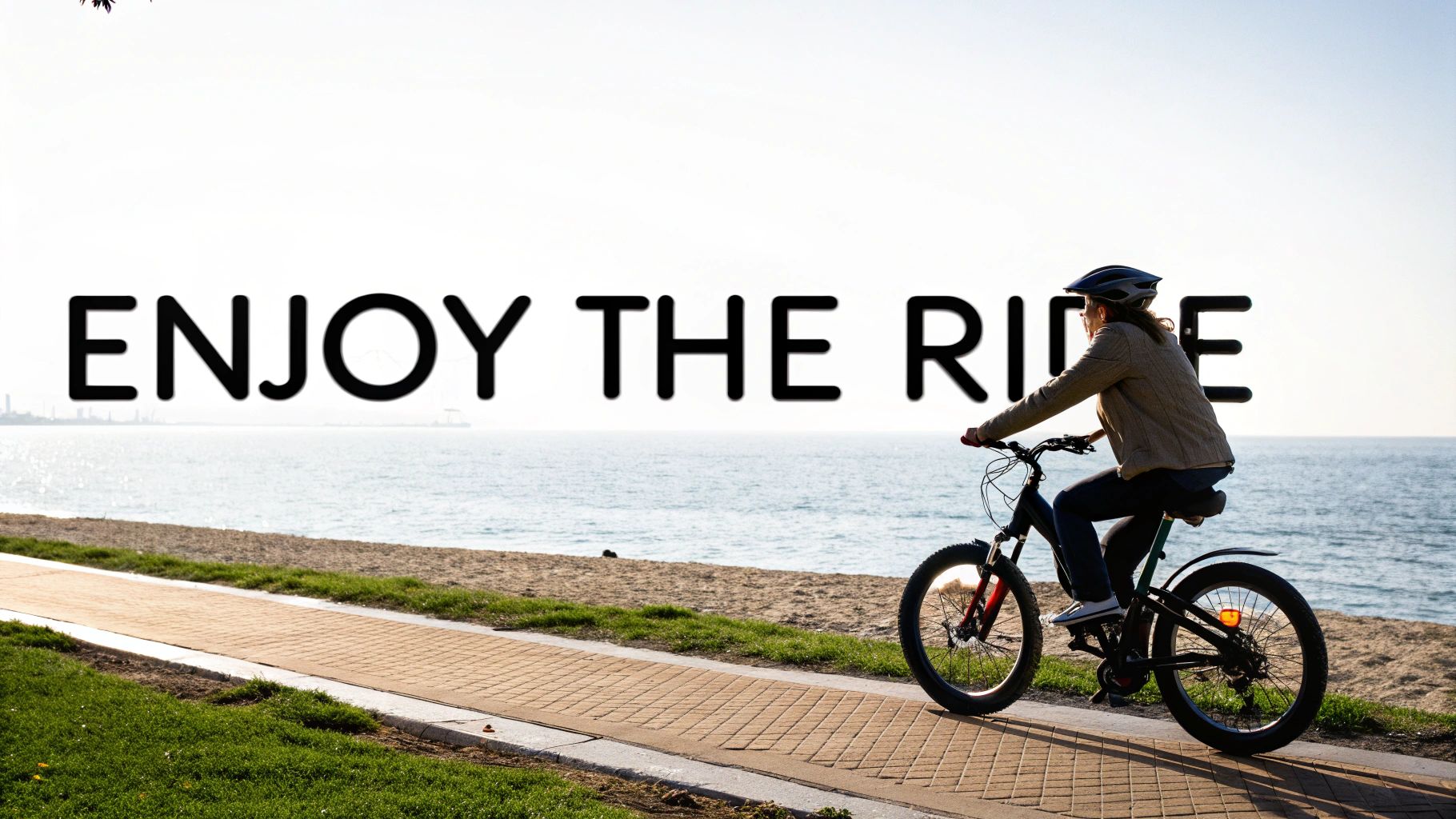 Osoba w kasku jedzie na rowerze przybrzeżną ścieżką nad oceanem, z nałożonym na nią napisem „CIESZ SIĘ JAZDĄ”.