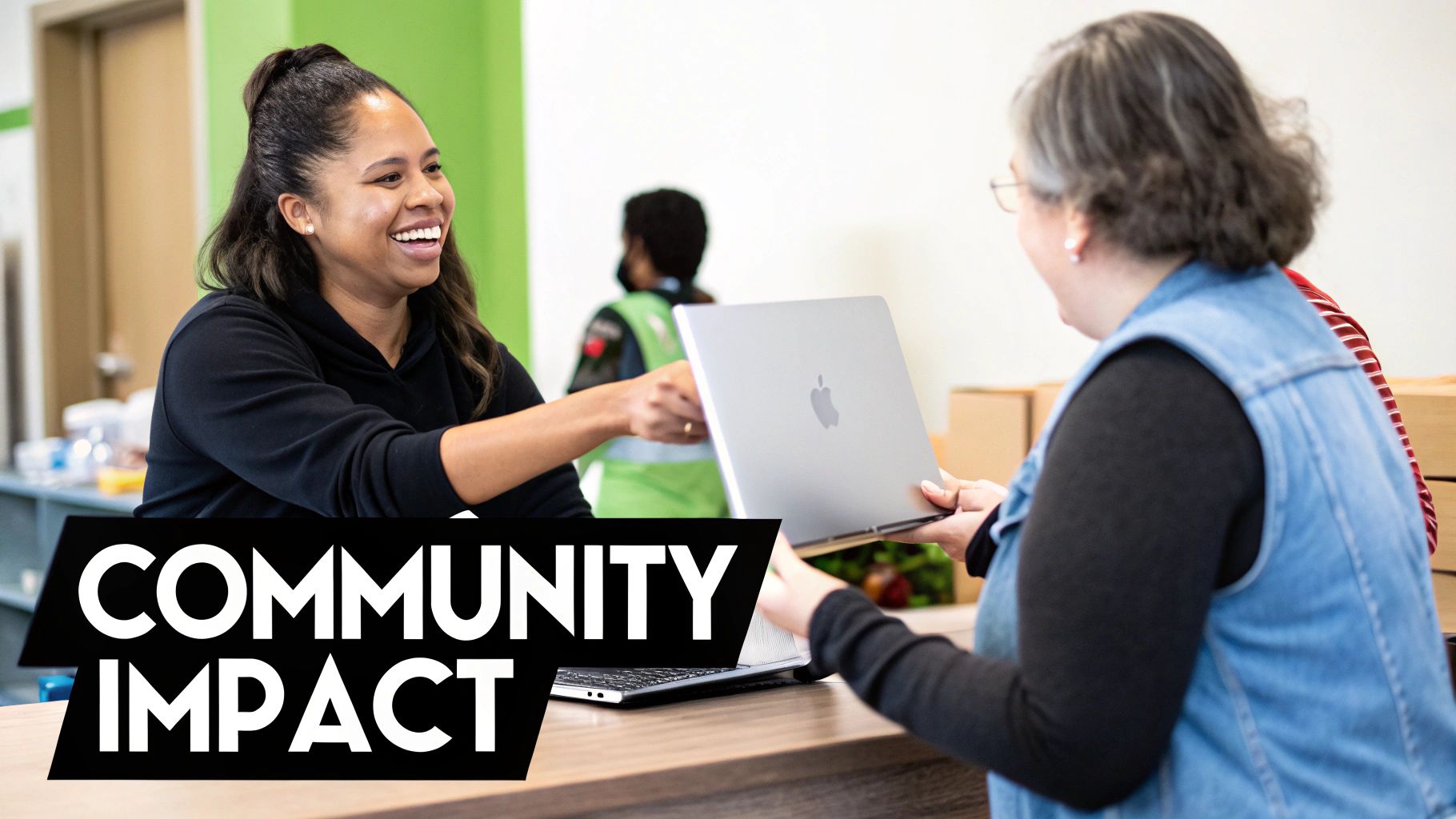 A smiling woman hands a silver laptop to another woman at a desk, showcasing community impact.