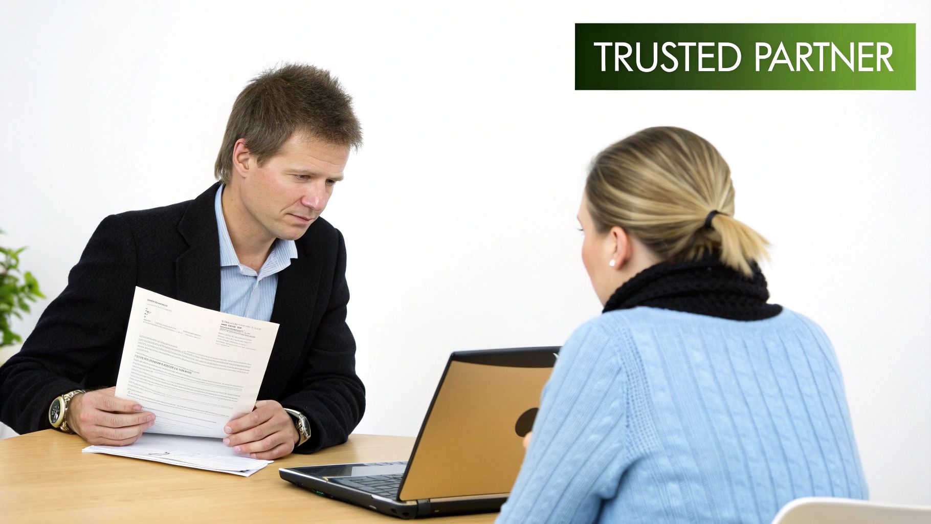 A man and woman are seated at a table during a business meeting, reviewing documents and a laptop.