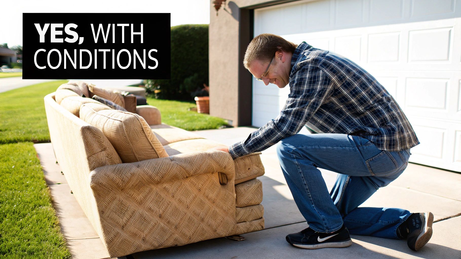 A man in a plaid shirt inspects an old, tan sofa outdoors on a driveway.