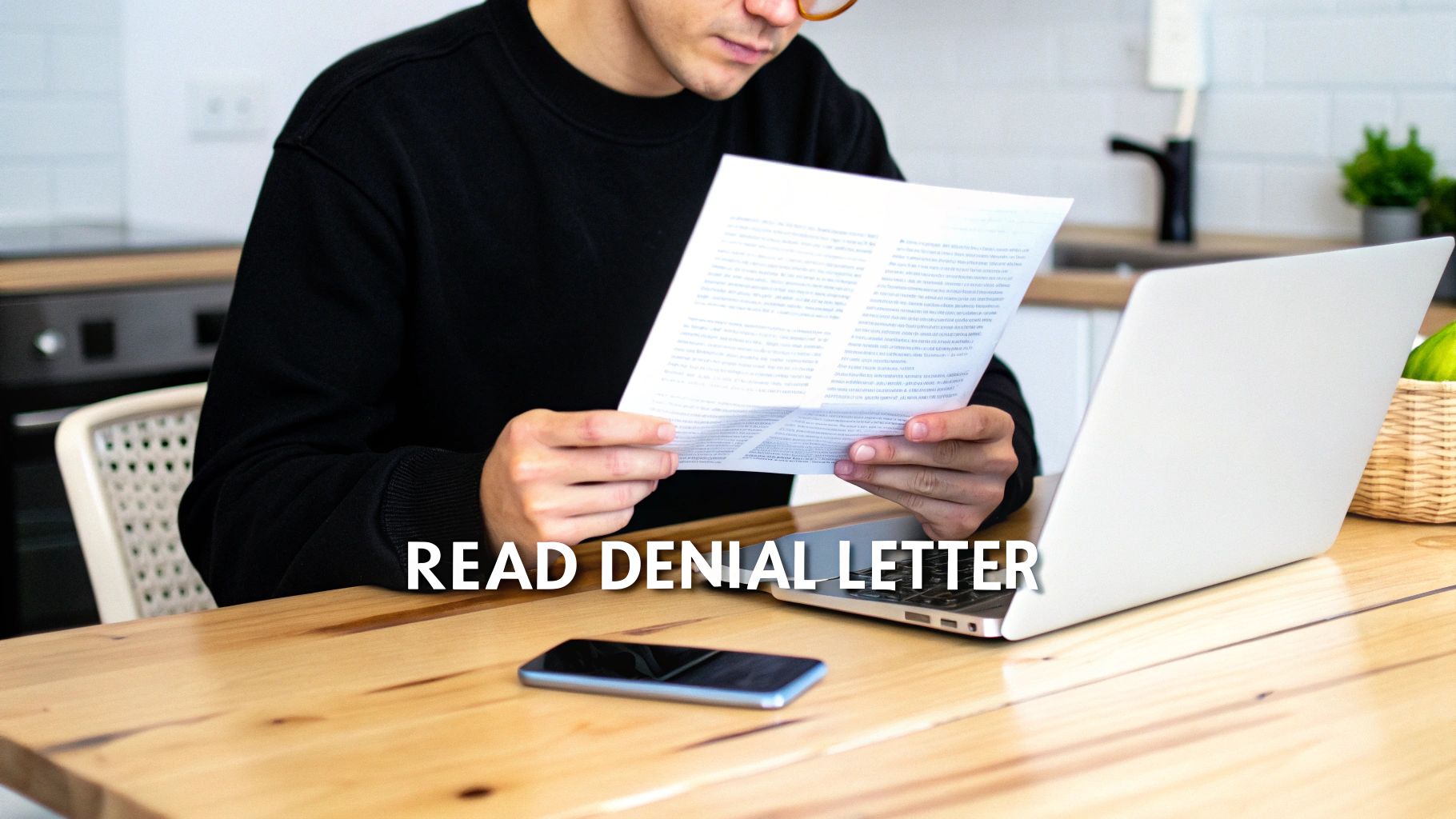 A person in a black sweater and glasses reads a denial letter at a wooden desk with a laptop.
