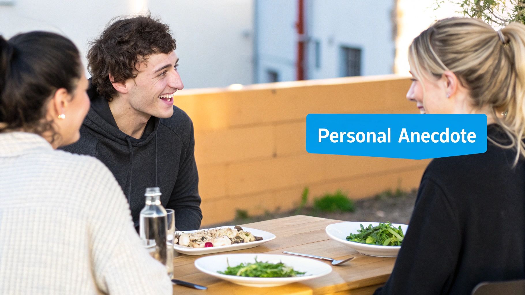 Three friends laughing and talking around a wooden table with food and drinks outdoors.