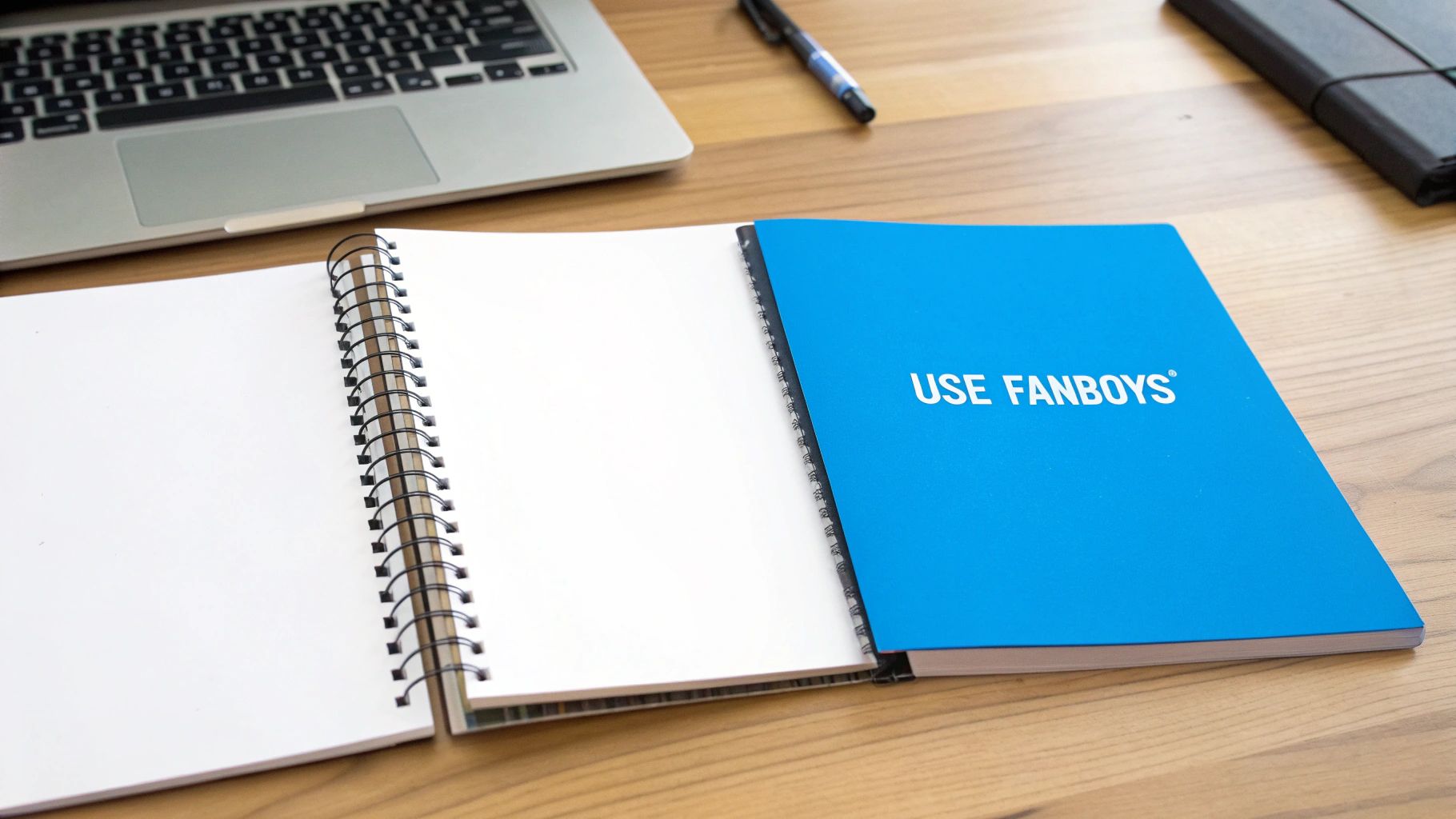 A wooden desk with an open spiral notebook showing blank pages, a blue "USE FANBOYS®" notebook, a laptop, and a pen.