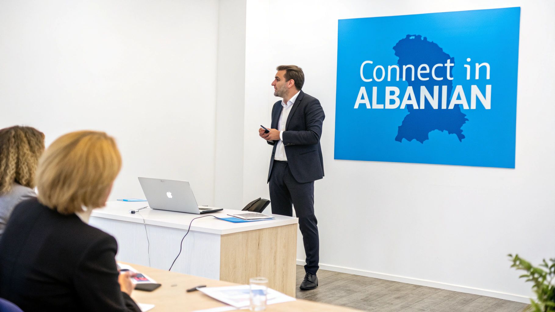 A man in a suit presents to two women in a modern room with a 'Connect in ALBANIAN' sign.