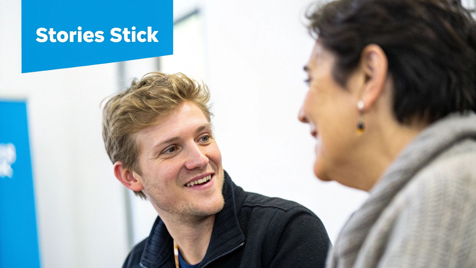 A smiling young man in a black jacket engaging in conversation with an older woman, with a blue banner that says "Stories Stick".