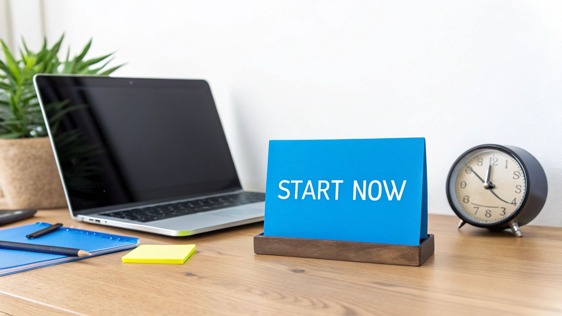 A clean and organized desk workspace featuring a laptop, plant, clock, and a 'START NOW' sign.