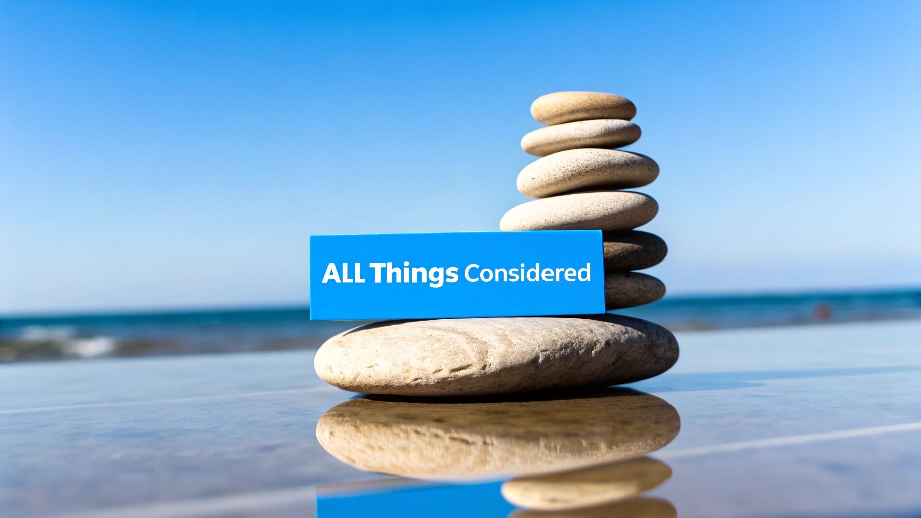 Blue sign 'ALL Things Considered' in front of balanced zen stones by the sea, reflecting on calm water.