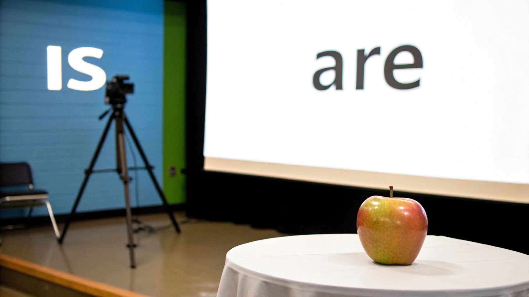 A red and green apple on a table, with screens displaying 'IS' and 'are' in the background.