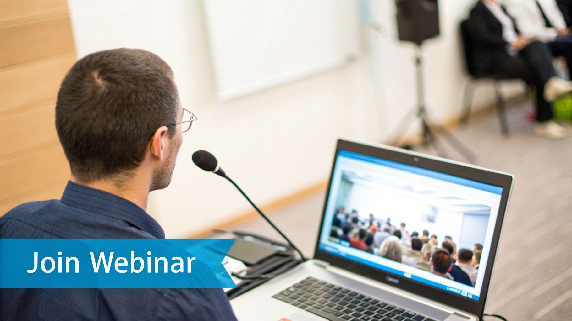 A man with short hair and glasses attends an online webinar, speaking into a microphone.