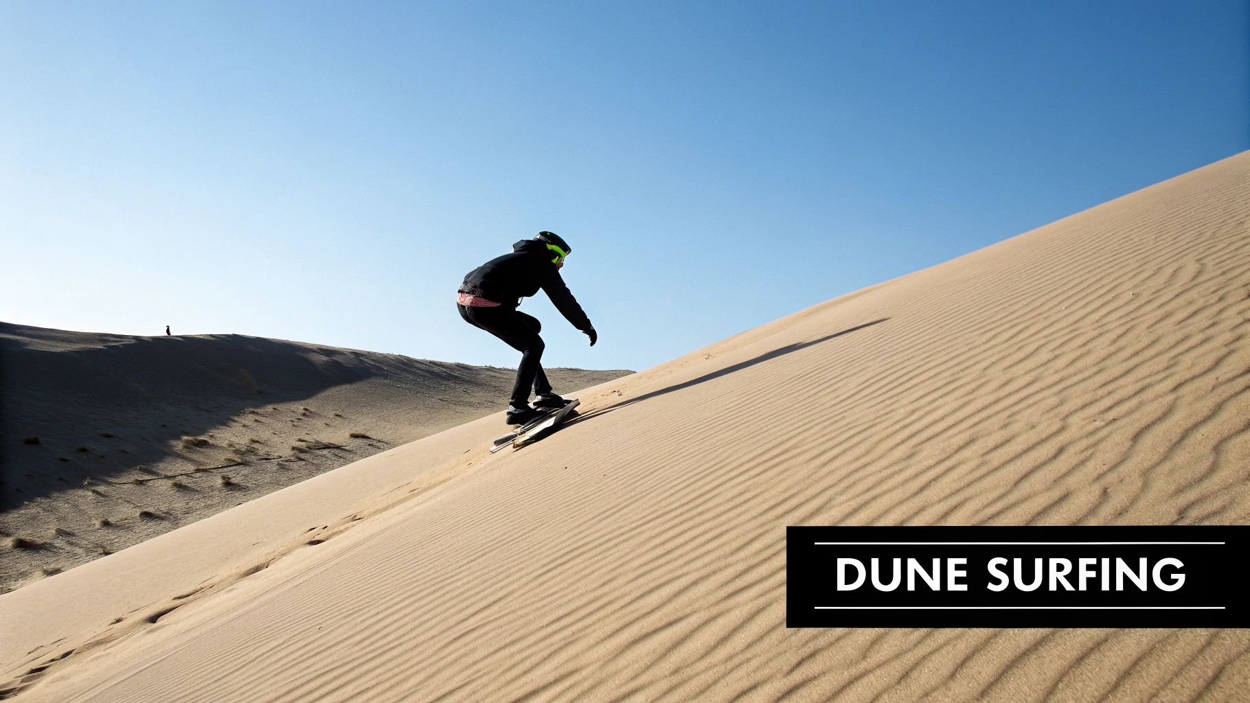 A person in black clothing and goggles sandboards down a large, striped sand dune under a clear blue sky.