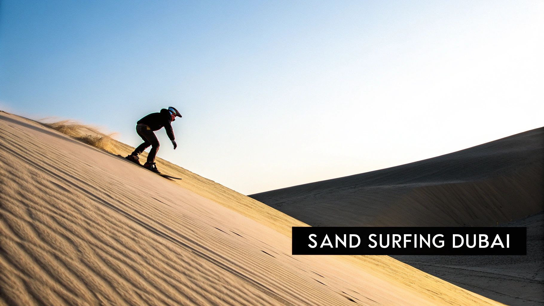 A person sandboarding down a large desert dune, kicking up sand, under a clear blue sky.
