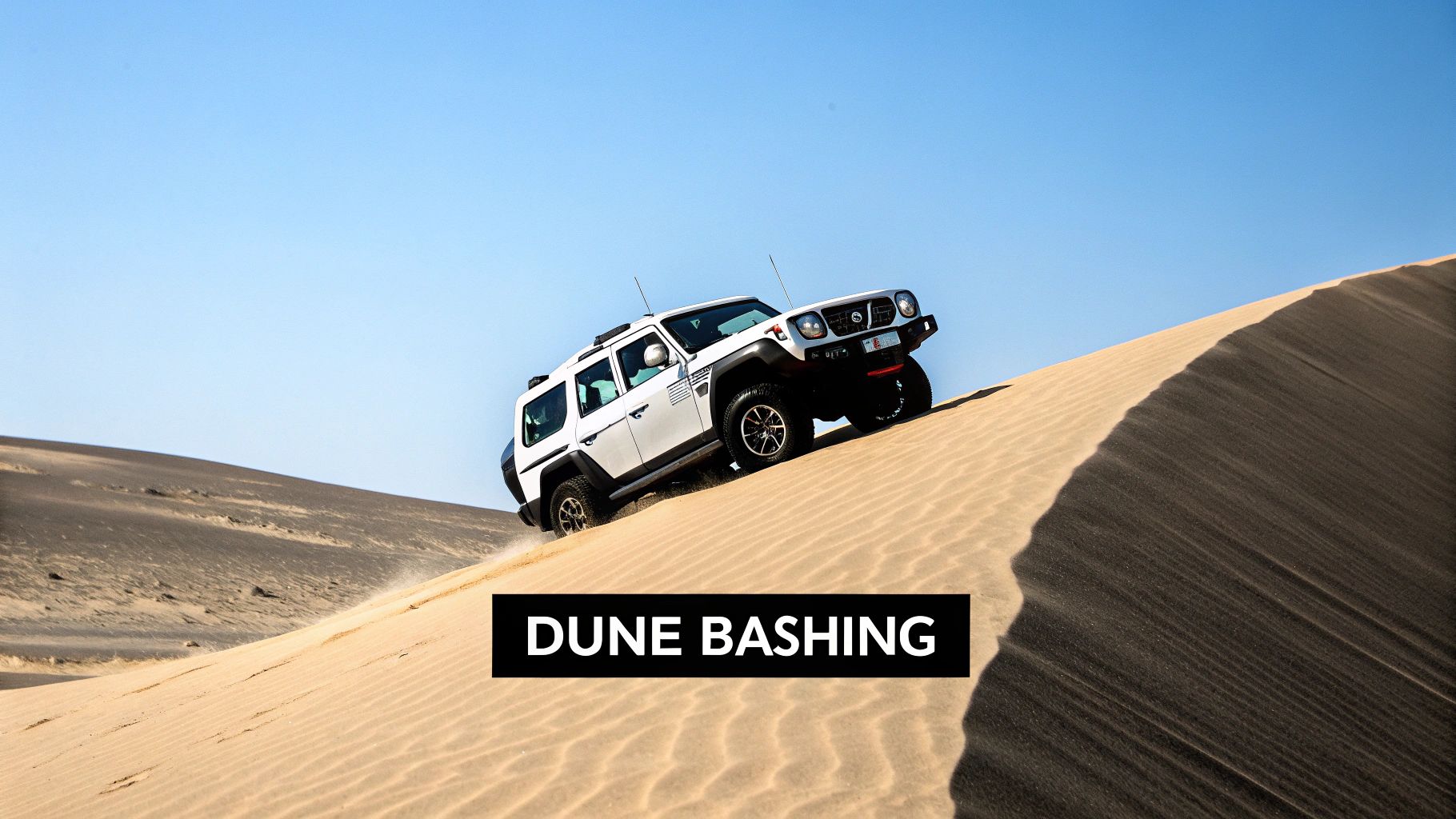 A white SUV ascends a steep sand dune, kicking up sand, under a bright blue sky, showcasing dune bashing.