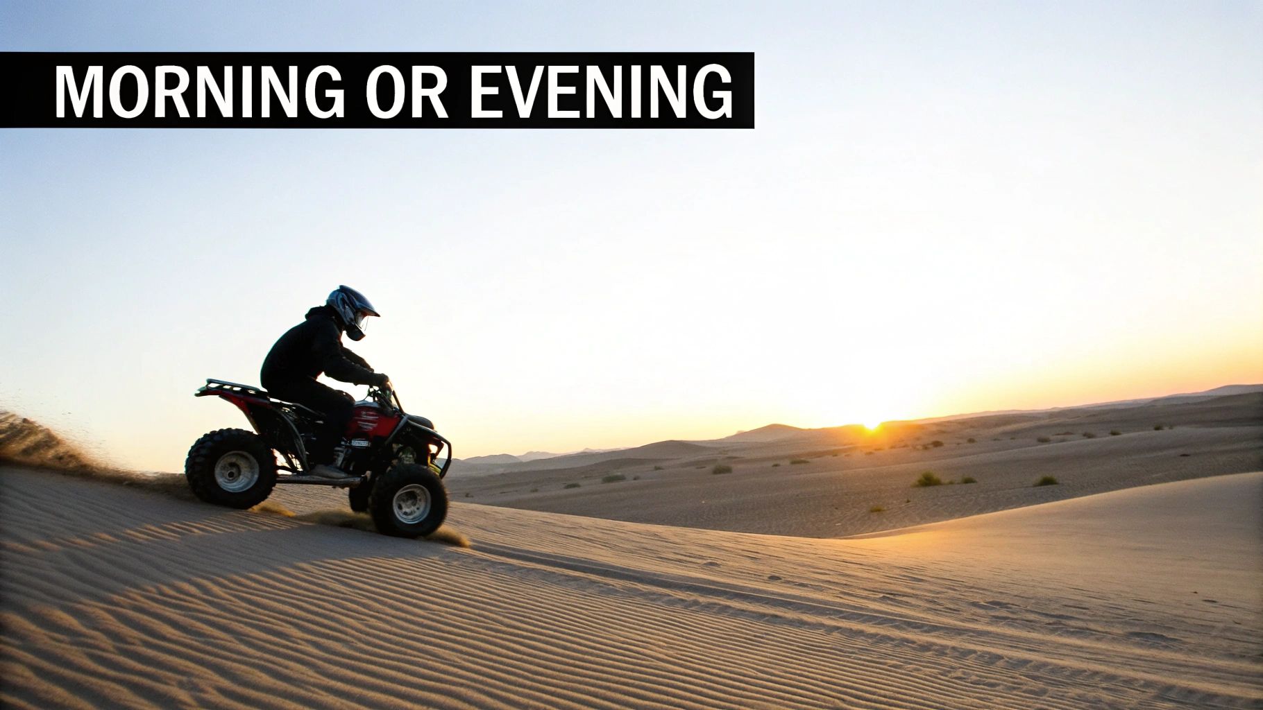 A person in a helmet riding a quad bike up a desert sand dune with the sun low on the horizon.