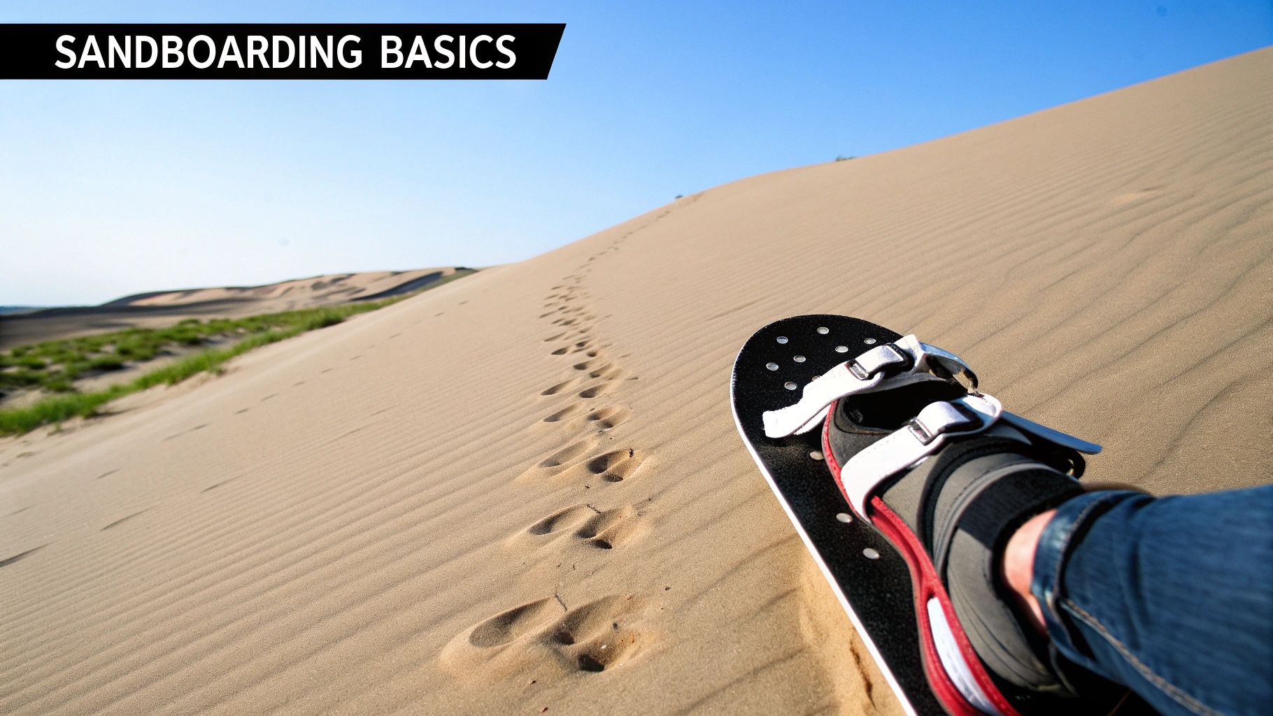 A sandboarder's foot on a sandboard, ready to descend a vast sandy dune with footprints under a clear sky.
